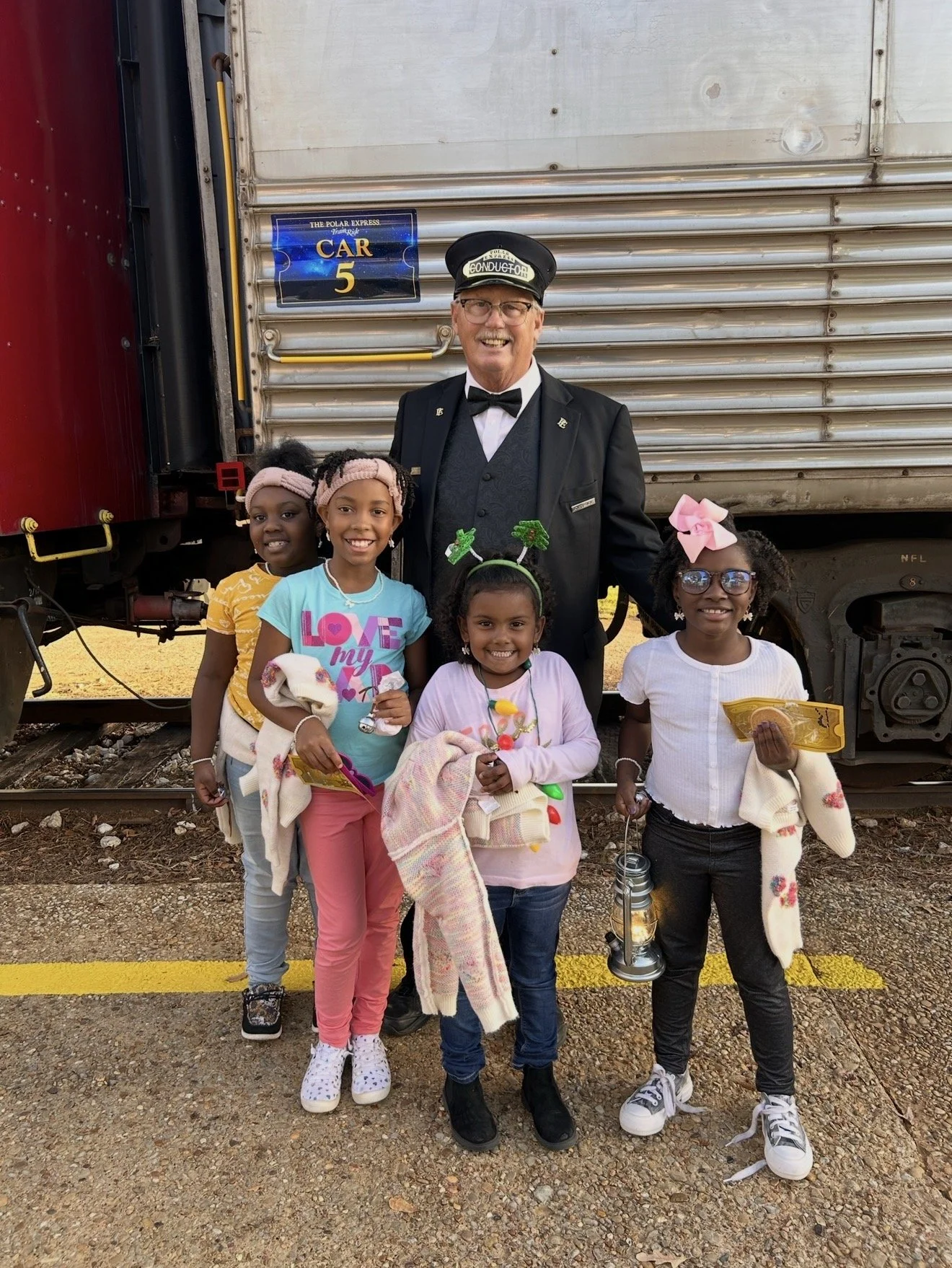 Group of five young girls and a man dressed as a train conductor posing outside next to a train car. The girls are smiling and holding various items, and are wearing colorful outfits and accessories. The man is smiling and dressed in a train conductor's uniform with a hat that says "Conductor."