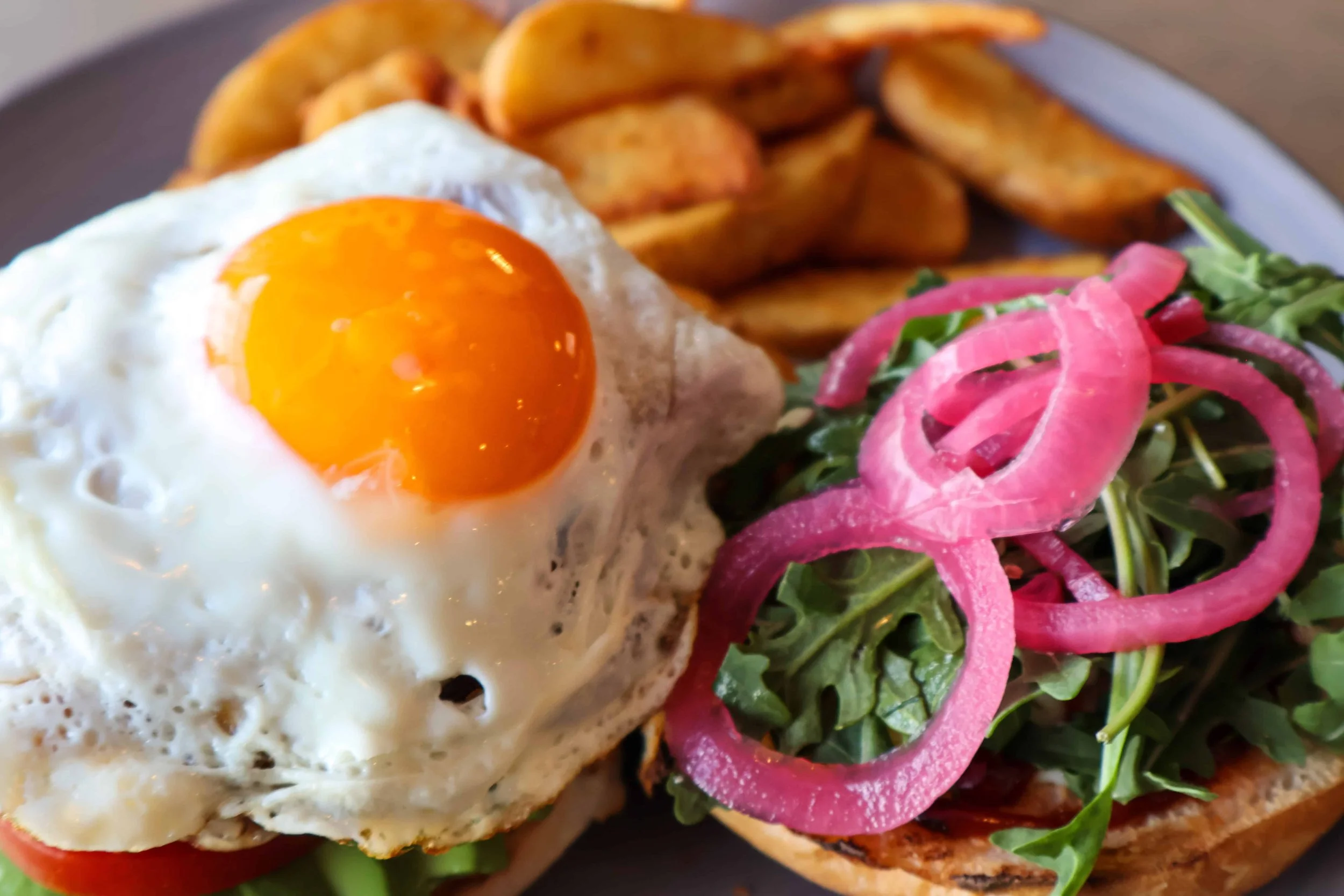 A plate with an open-faced sandwich featuring arugula, pink pickled onions, grilled bread, a fried egg with a runny yolk, and potato wedges in the background.
