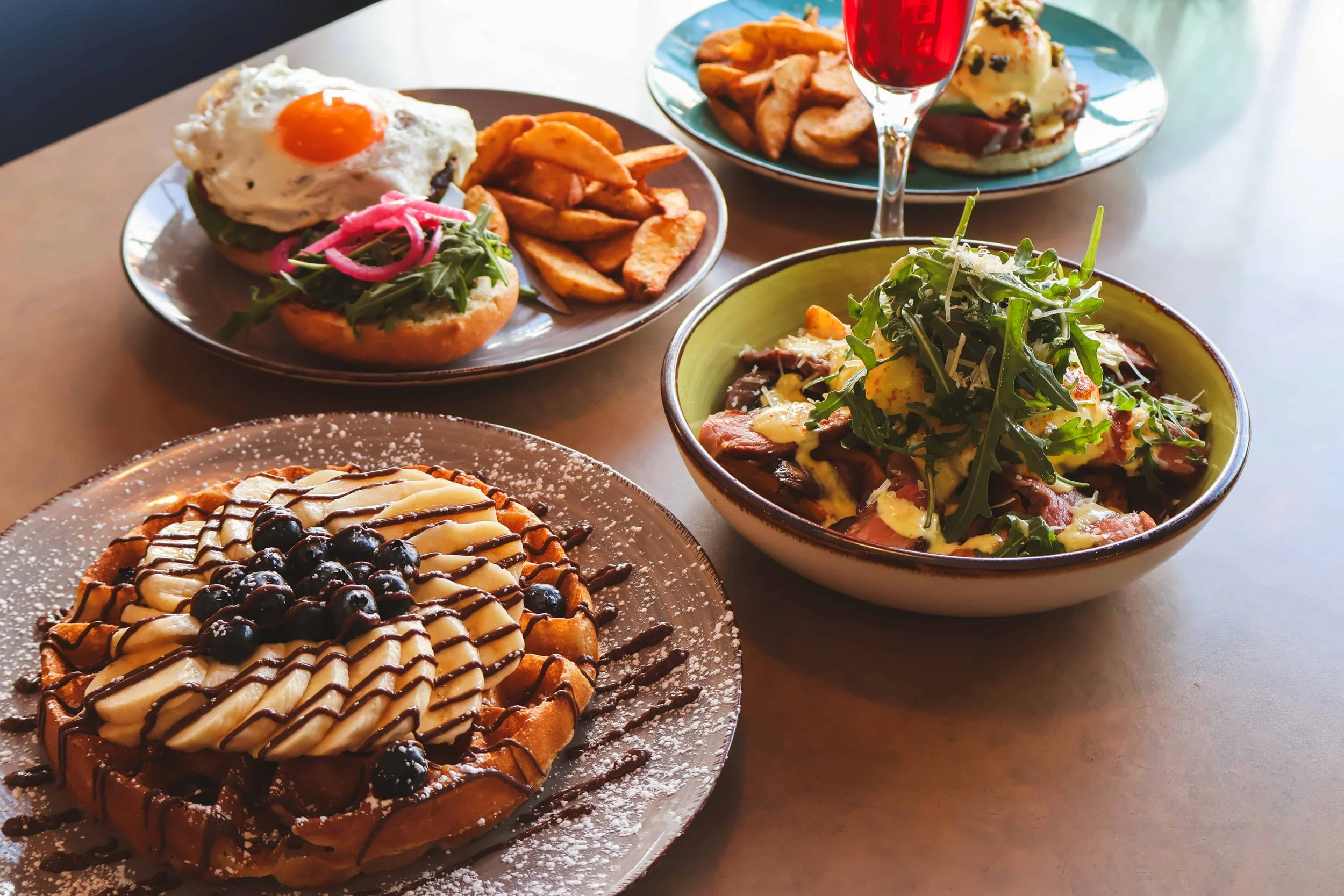 A table with various dishes including a Belgian waffle topped with bananas, blueberries, chocolate drizzle, and powdered sugar, a burger with a fried egg, pickles, and lettuce, a bowl of salad with greens and dressing, a plate of potato wedges, and an open-faced sandwich with toppings, accompanied by a glass of red drink.