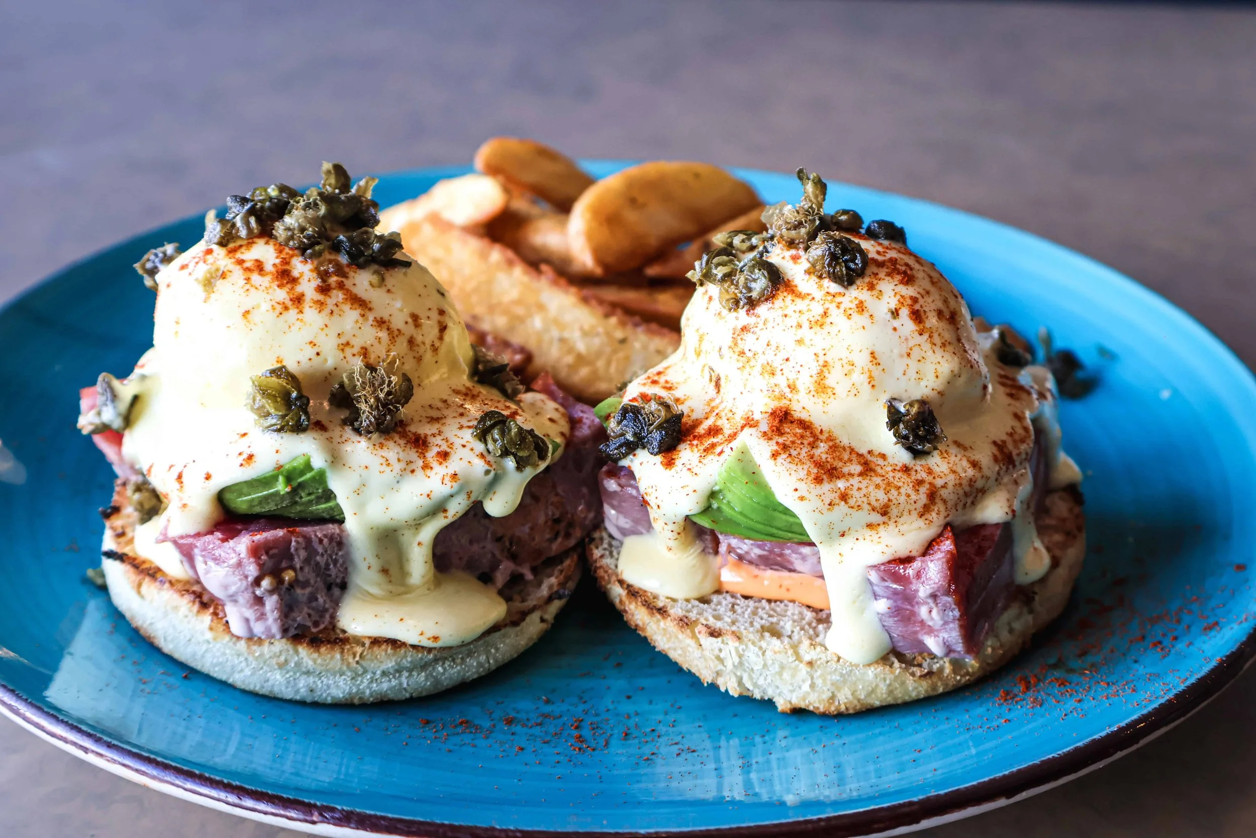 Two eggs benedict with hollandaise sauce, chopped herbs, and seasoning, served on toasted English muffins with avocado and ham, accompanied by roasted potatoes on a blue plate.