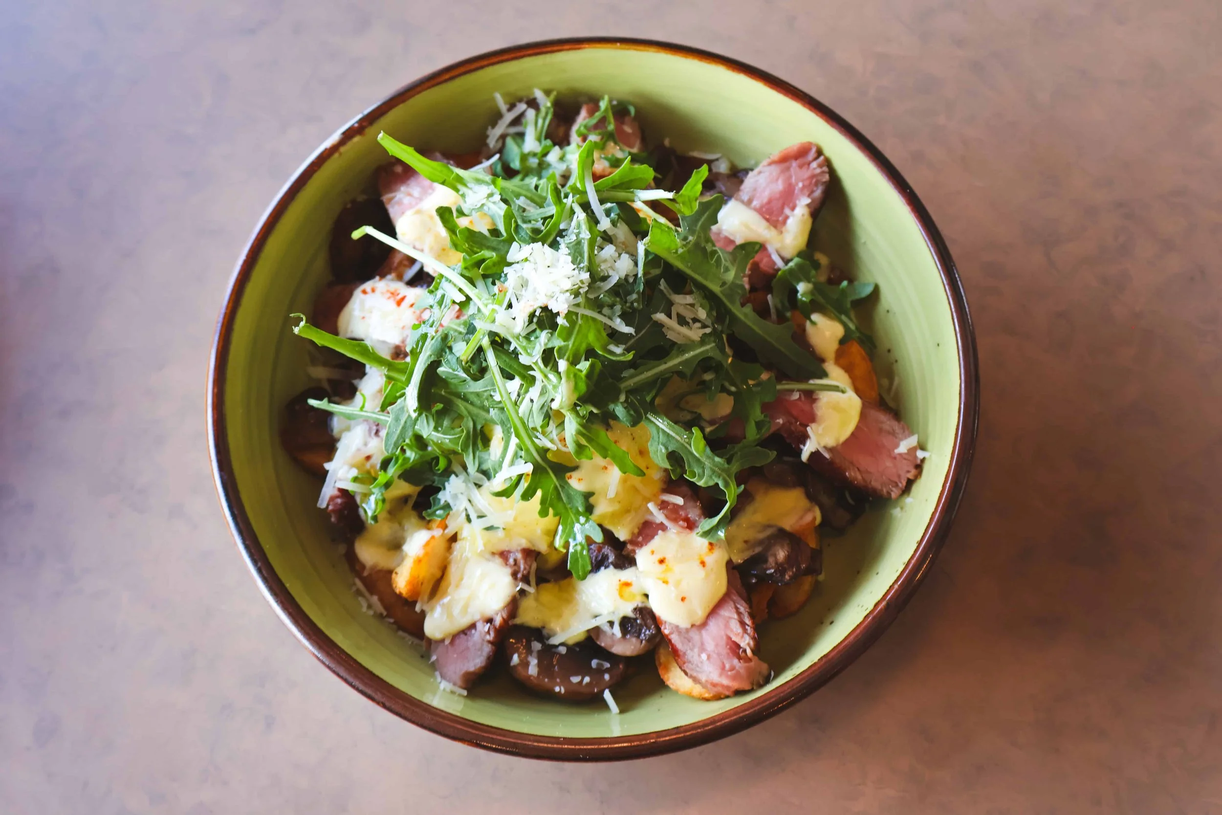 A bowl of salad with arugula, cheese, grilled steak slices, mushrooms, and shaved Parmesan cheese on top, on a wooden surface.