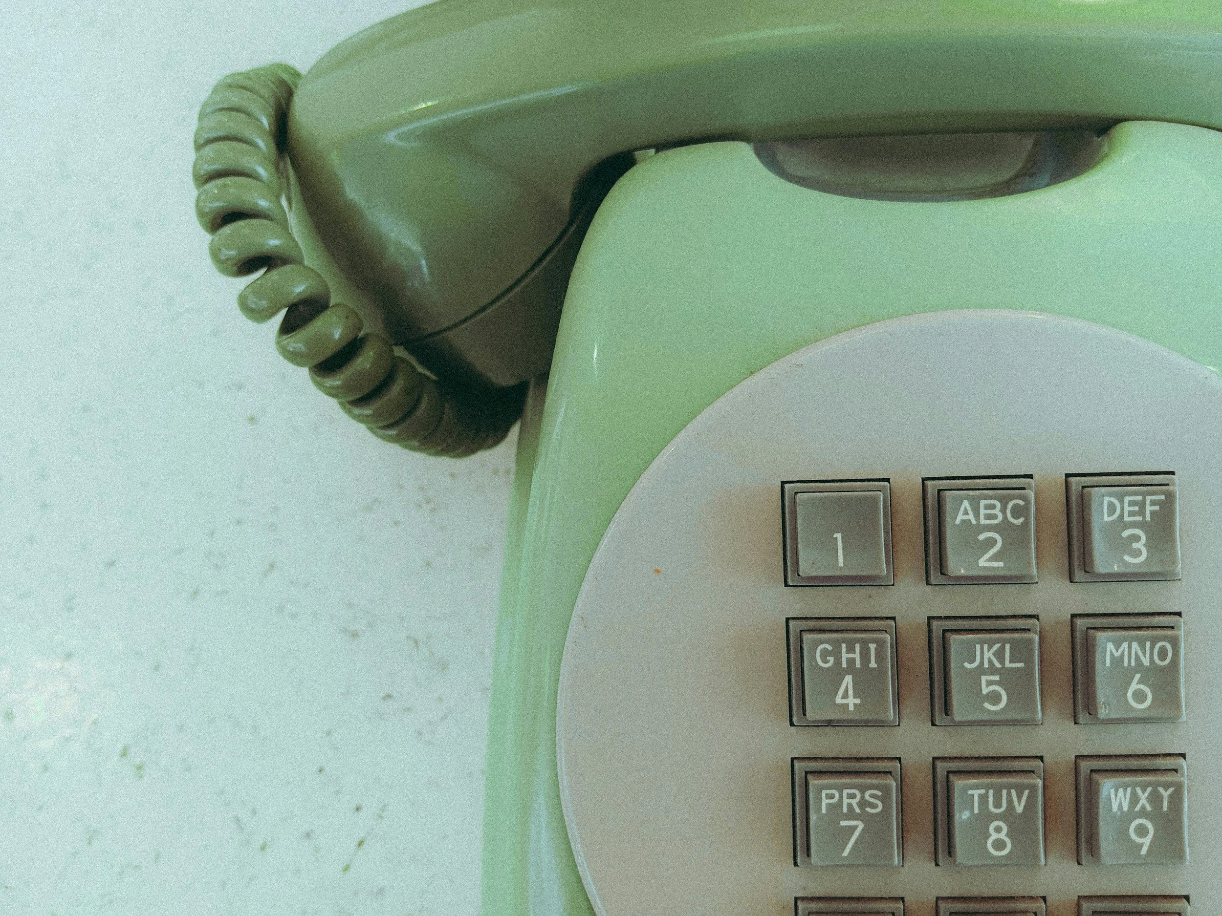 A close-up of a vintage rotary dial phone with a beige and light green color scheme, showing the rotary dial with numbered buttons and a coiled cord.