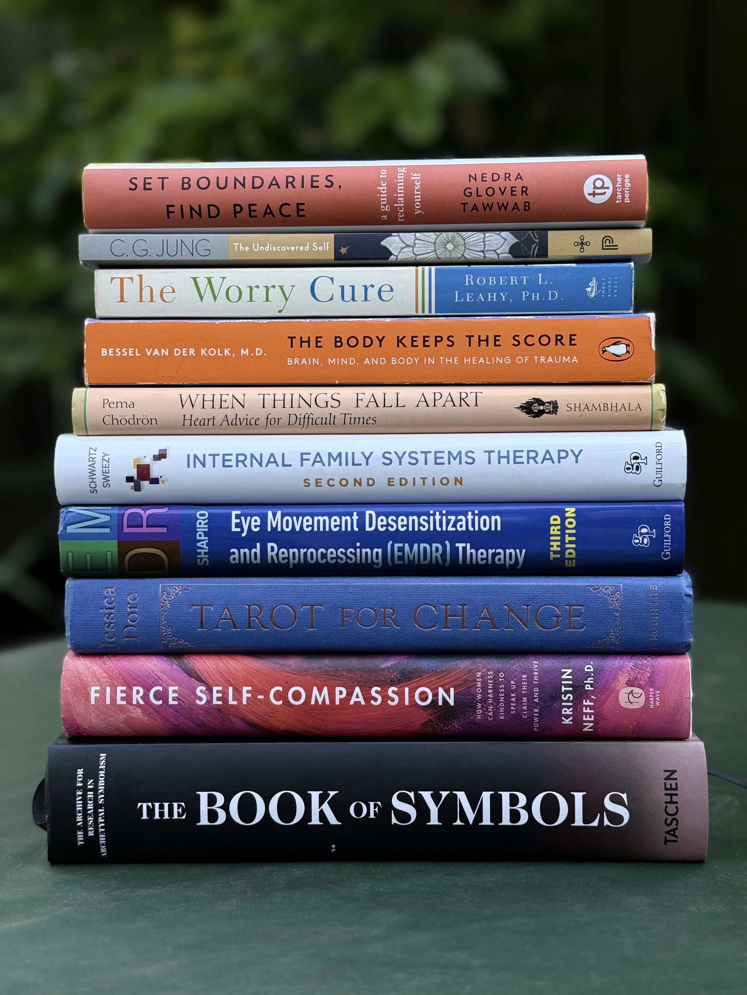 Stack of ten books on a dark green surface, with blurred green foliage in the background.