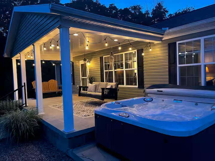 Cozy backyard patio with string lights, a cushioned sofa, a swing, and a hot tub at dusk.