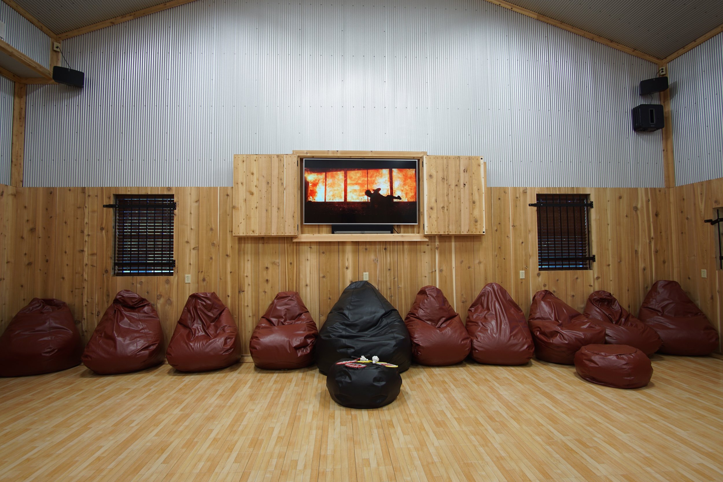 A room with wooden walls and floor, featuring a large TV displaying a scene from a fire, surrounded by bean bags in front of the wall.