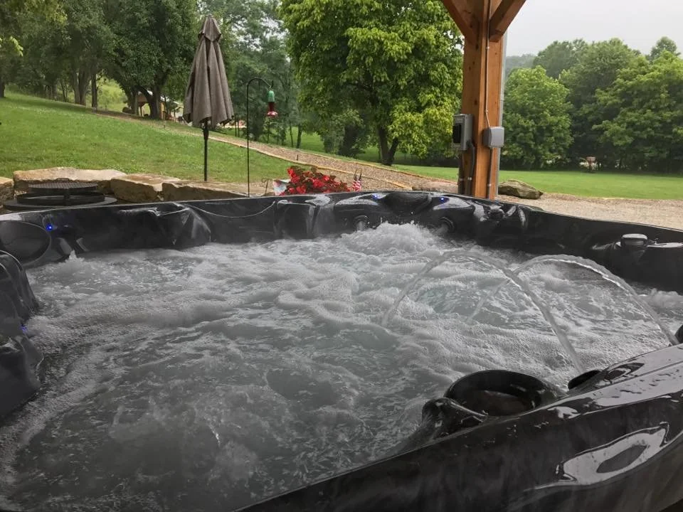 A hot tub with bubbling water outdoors on a covered patio, with trees and grass in the background.