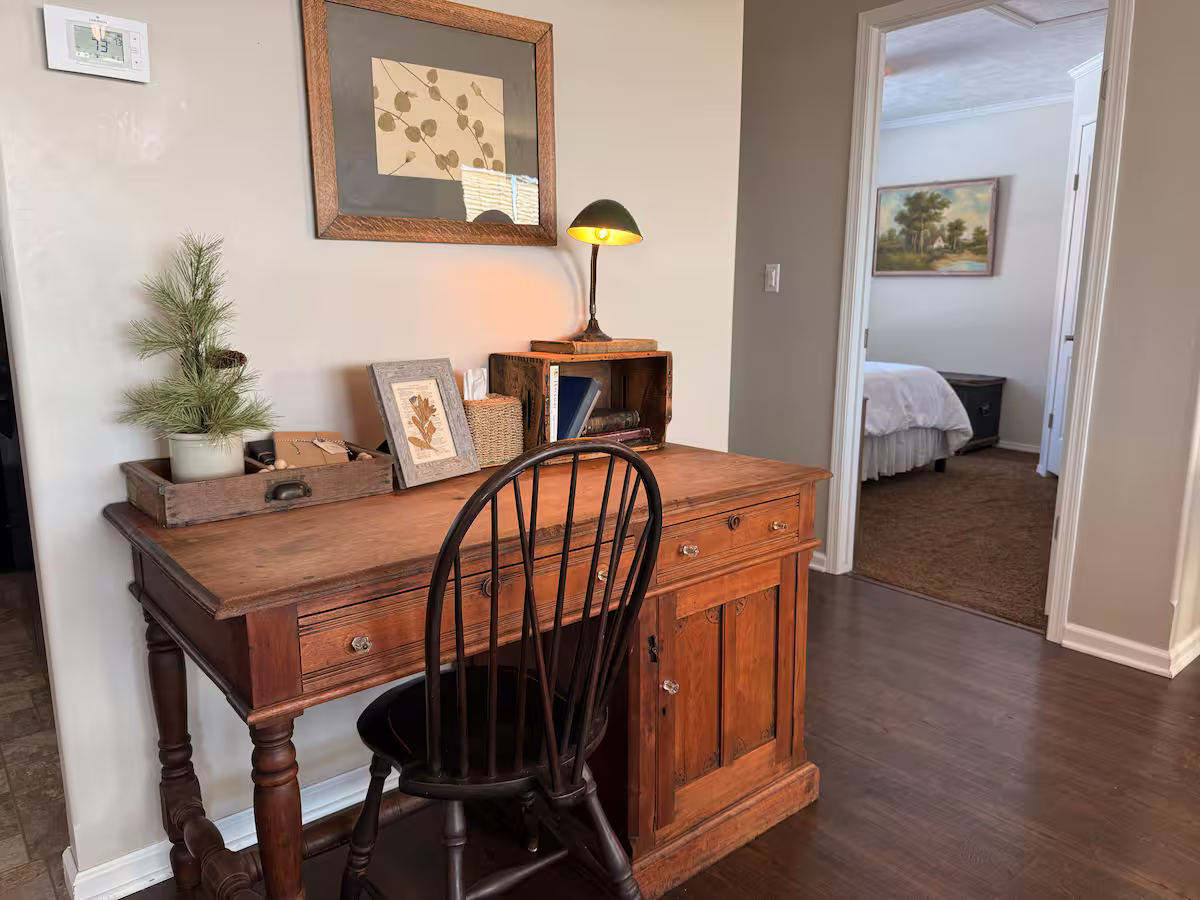 A wooden desk with a black chair in front of it, decorated with a small potted pine tree, framed artwork, and a tray with miscellaneous items. A small table lamp with a green shade illuminates part of the desk. Behind the desk is a wall with a framed