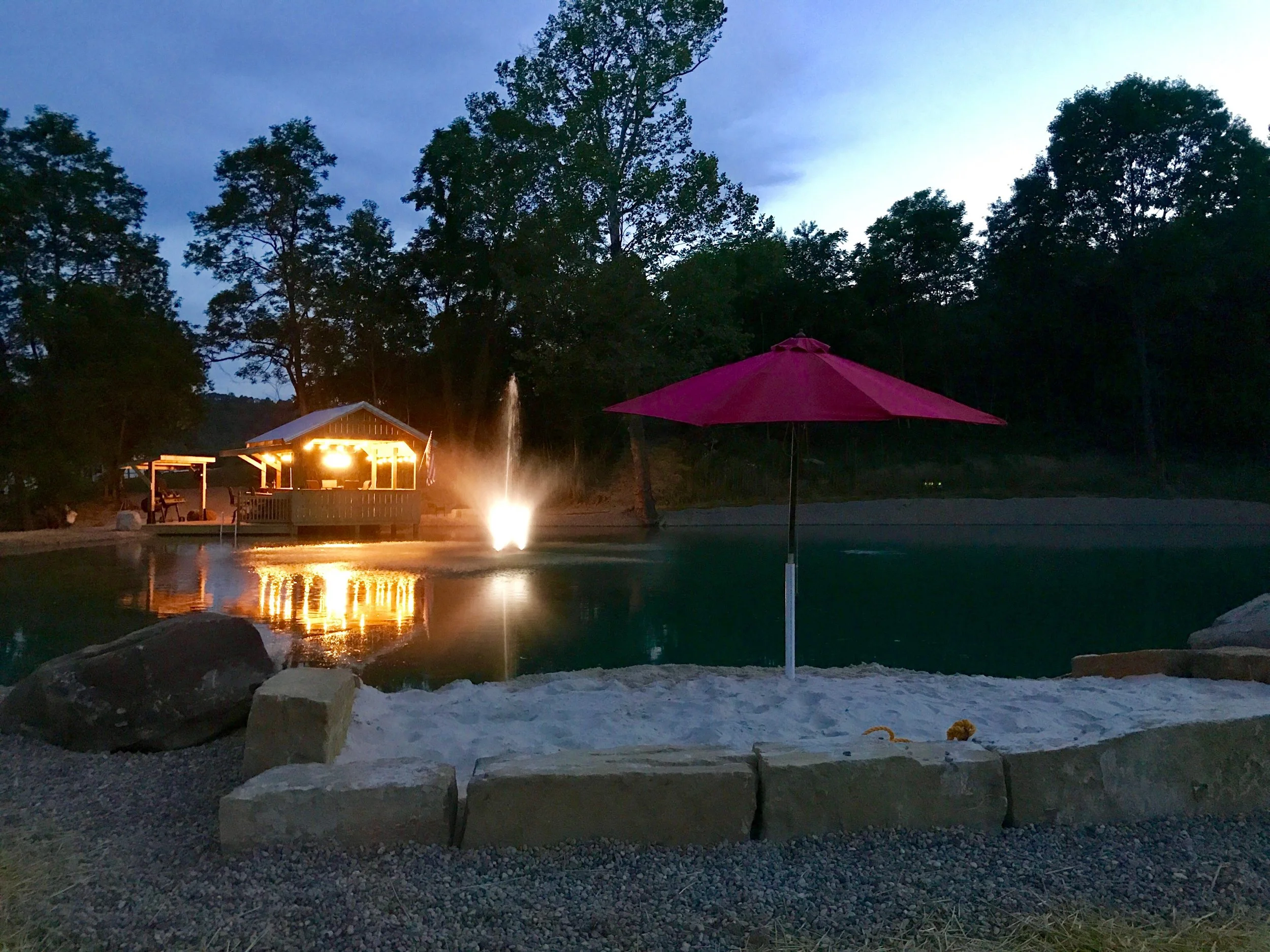 Nighttime scene at a backyard pool with a pink umbrella in the foreground, a water fountain in the middle, illuminated house in the background, and surrounding trees.