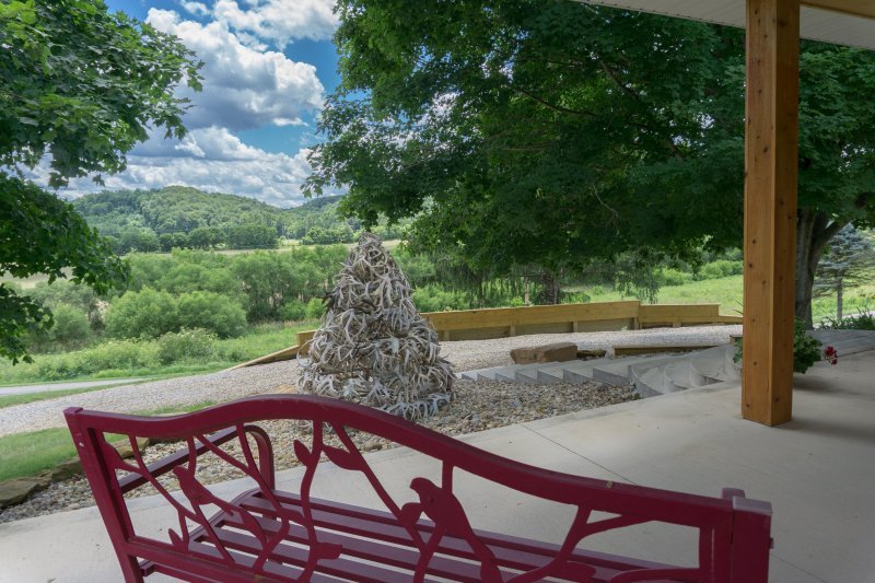 View from a porch showing a red metal bench, a tree with dense green foliage, a decorative tree sculpture made of intertwined branches, and a scenic landscape with hills and a cloudy sky in the background.