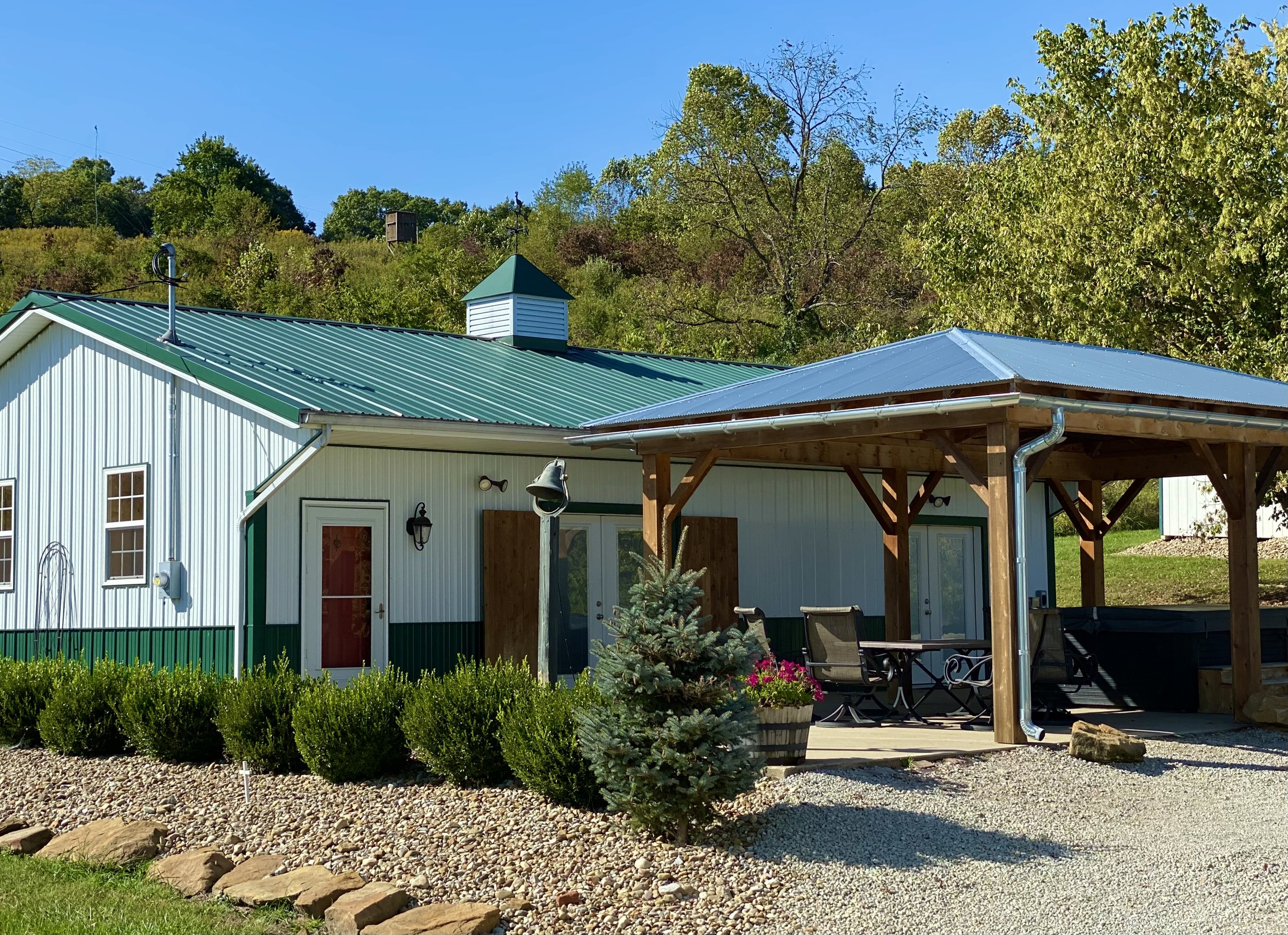 A building with a green metal roof and white vertical siding, featuring two windows and a door with red trim, and a small cupola on top. In front of the building, there is a covered patio area with outdoor furniture, including chairs and a table, and