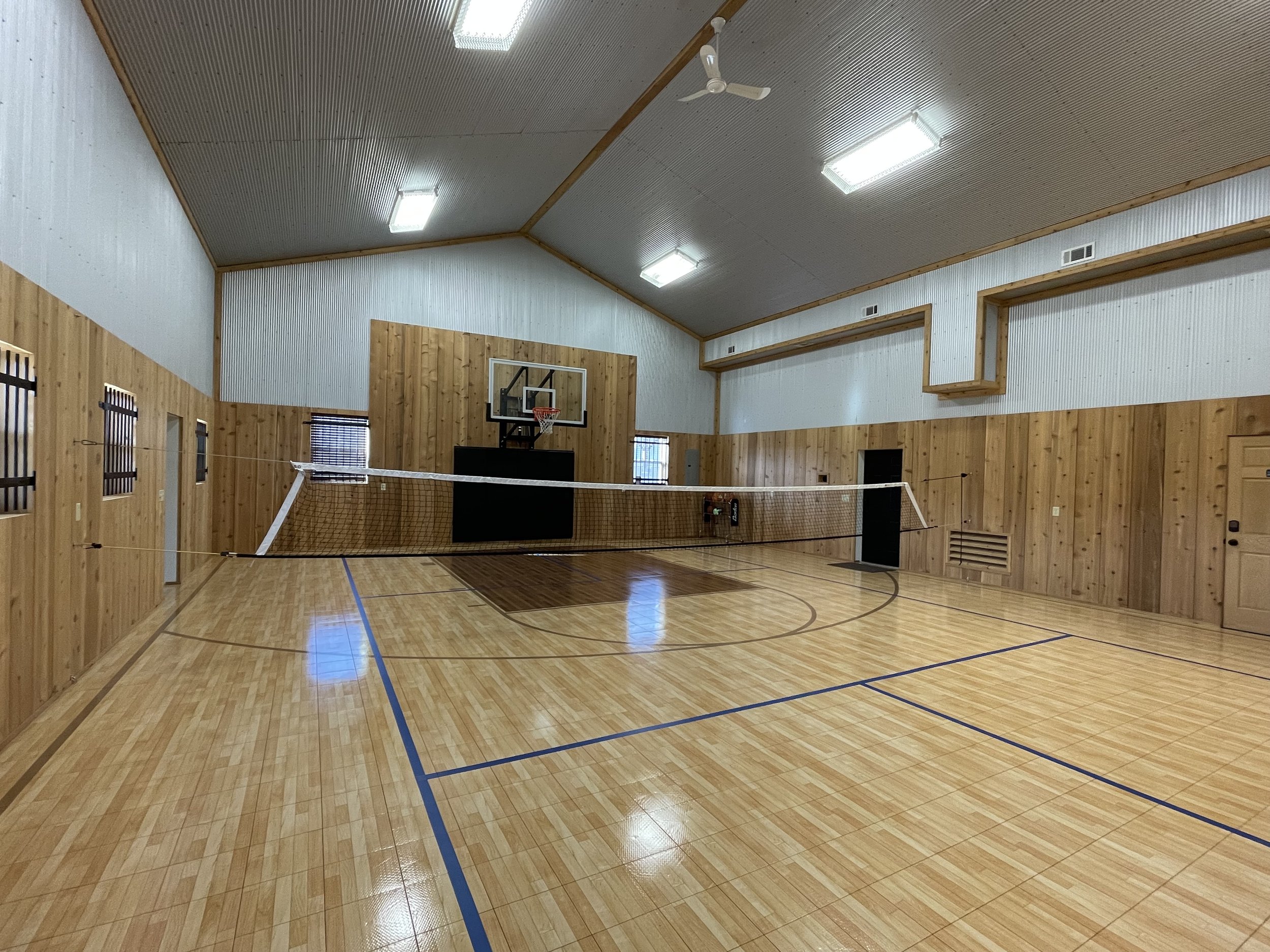 An indoor gymnasium with wooden walls and a wooden floor, featuring a volleyball net set up in the middle, a basketball hoop on the wall, and ceiling fans and lights.