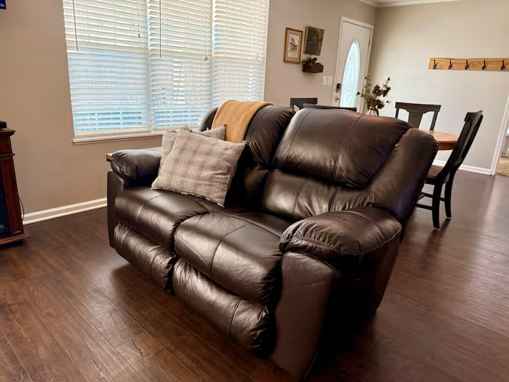 Living room with a black leather couch, a plaid cushion, and a yellow throw blanket. Behind the couch are white window blinds, and the room has hardwood floors and beige walls.