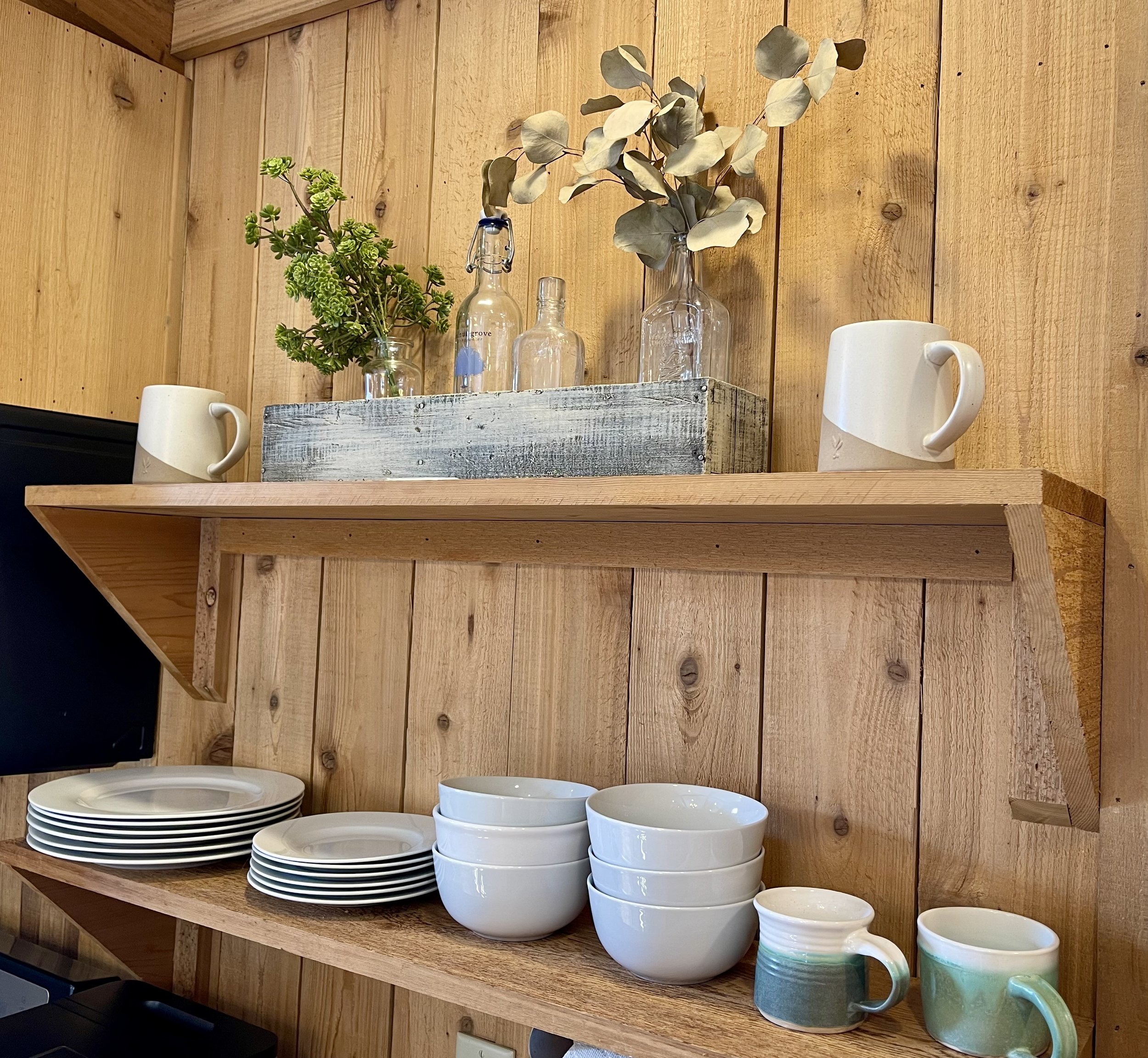 Wooden pantry shelf with white plates, bowls, and ceramic mugs, a rustic wooden box with glass bottles and plant arrangements.