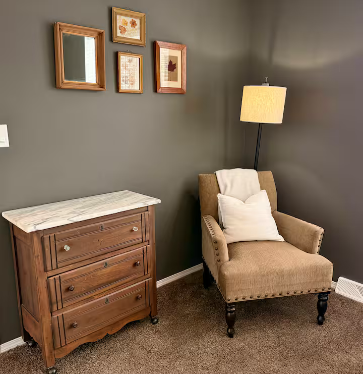 A cozy corner with a wooden dresser with a marble top, a beige armchair with nailhead trim and white pillow, a tall floor lamp with a beige shade, gray wall with four framed artworks, and brown carpeted floor.