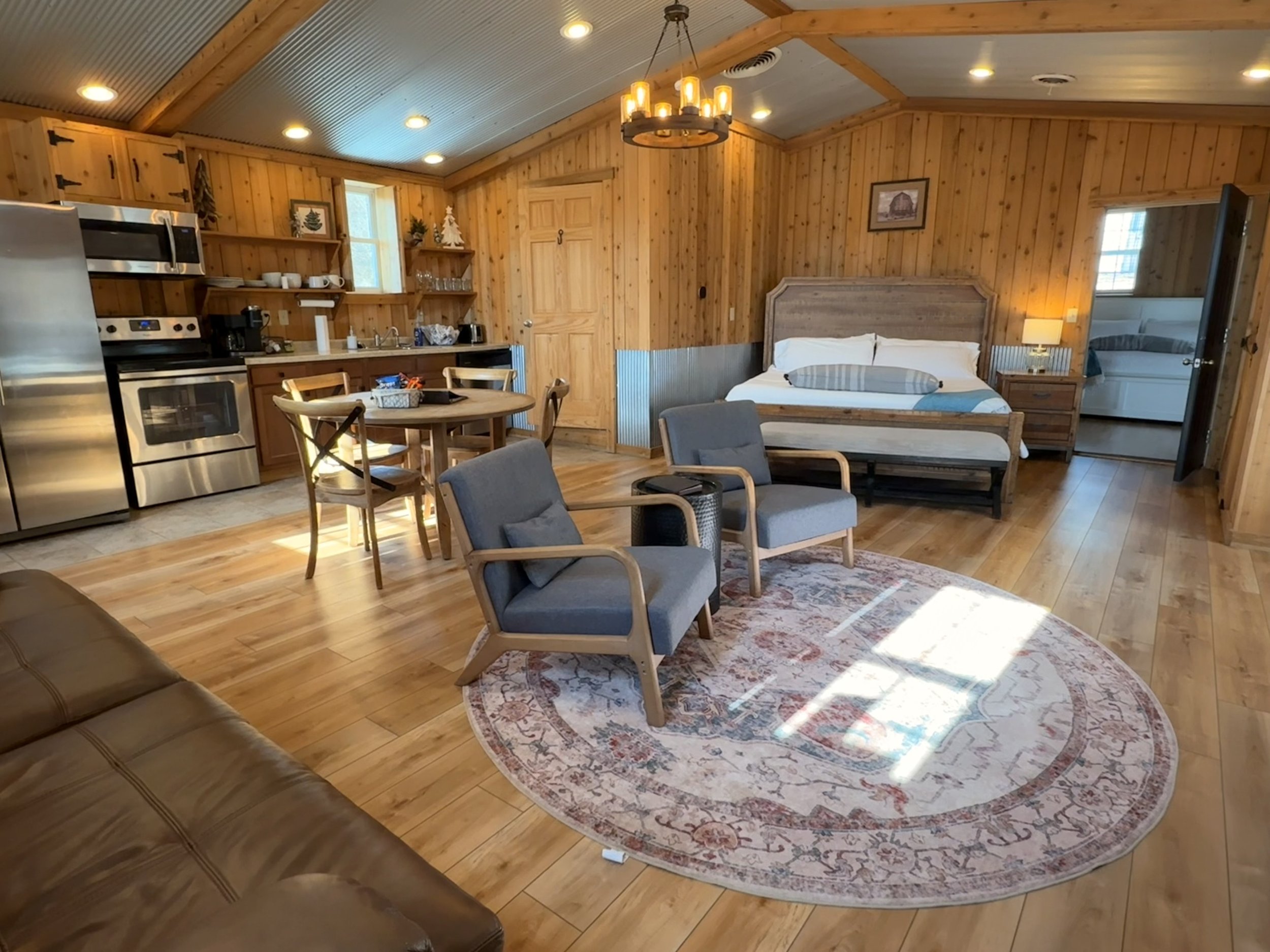 Open-concept living area with wooden walls and floors, includes kitchen, dining, and sleeping spaces, with seating area and bed near a window.