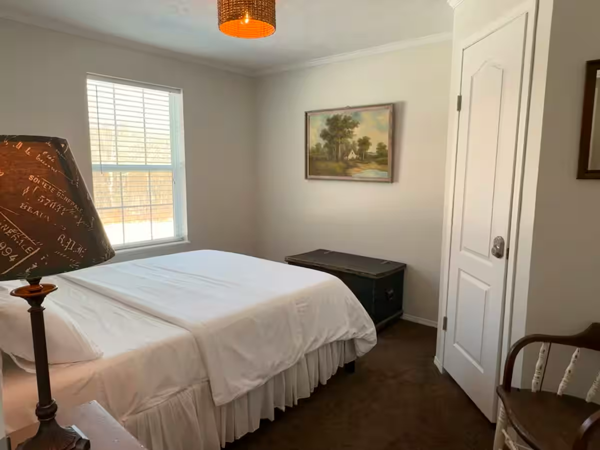 A bedroom with a bed, window, nightstand, framed picture on the wall, and a chair.