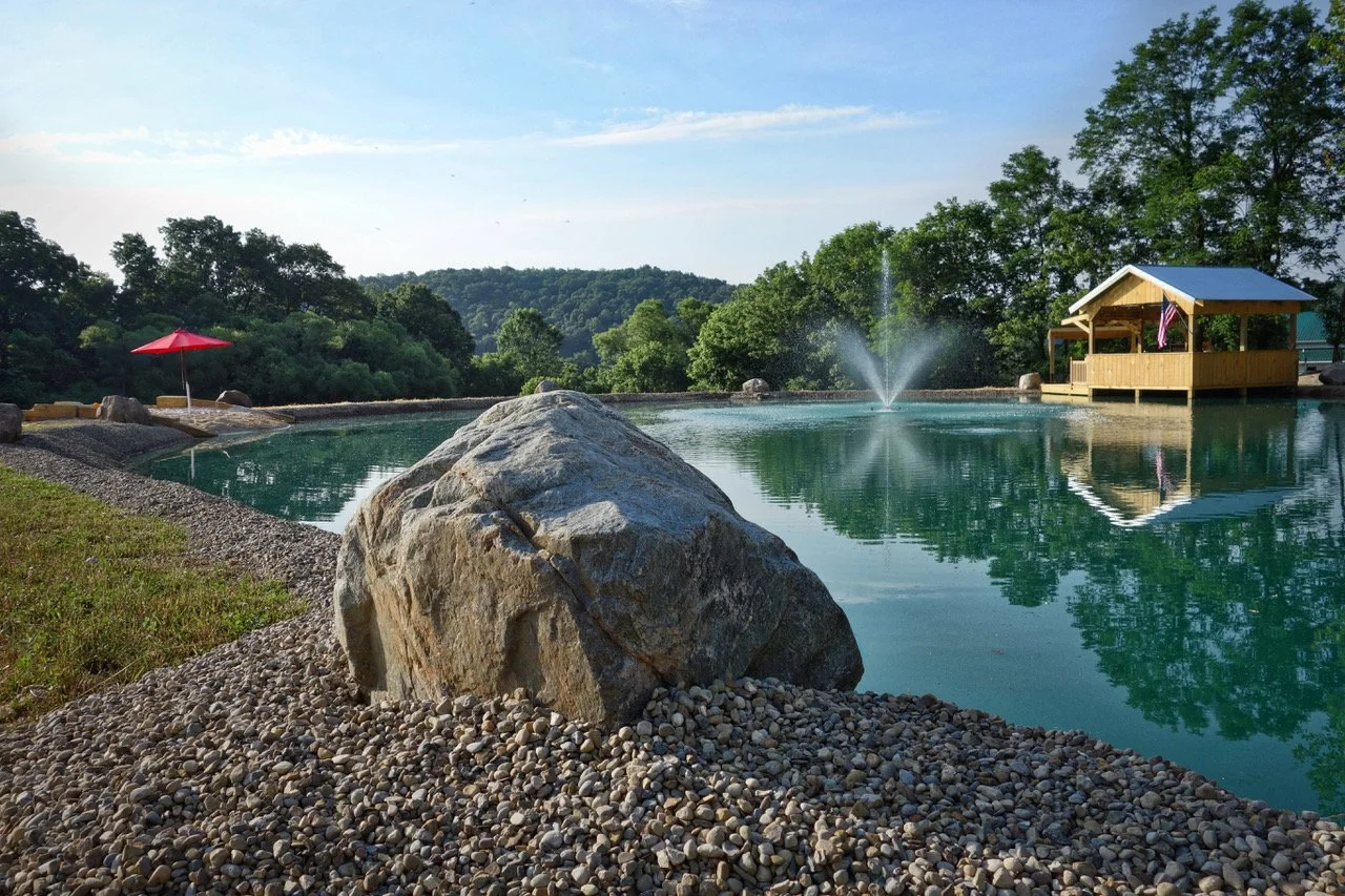 A peaceful outdoor scene with a small pond or pool, a large rock in the foreground, a red umbrella on the left, a fountain in the center, and a wooden pavilion with an American flag on the right, surrounded by green trees and hills under a partly clo