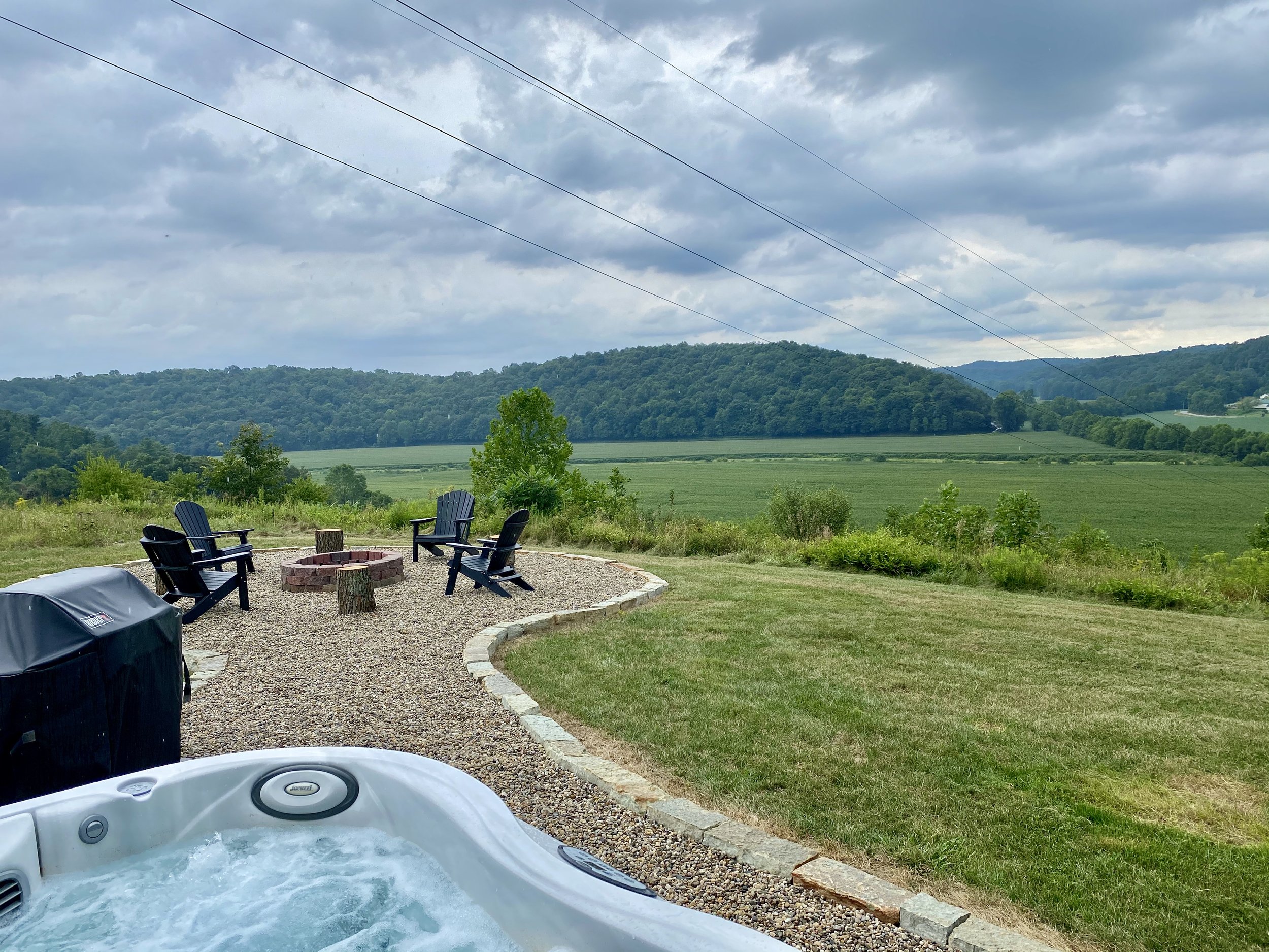 Hot tub with bubbling water in the foreground, outside patio area with four black chairs surrounding a fire pit, green lawn and trees, rolling hills and cloudy sky in the background.