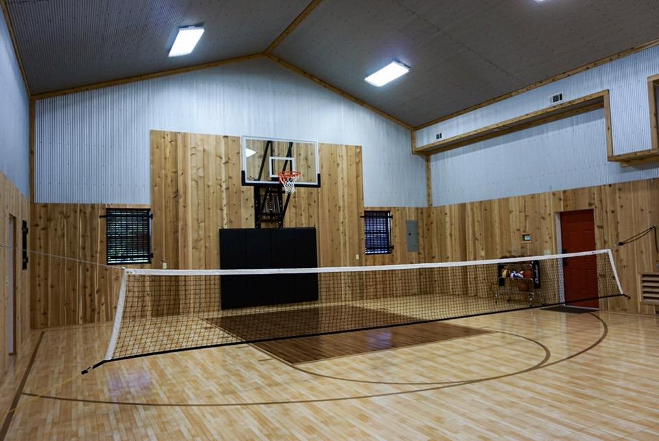 An indoor gymnasium with wooden walls and floor, featuring a volleyball net and a basketball hoop.