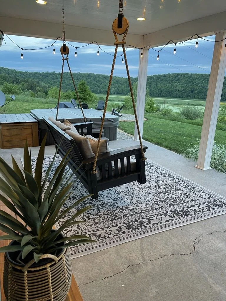 Covered porch with outdoor swing, rug, string lights, table and chairs, and a view of a grassy landscape with trees and hills.