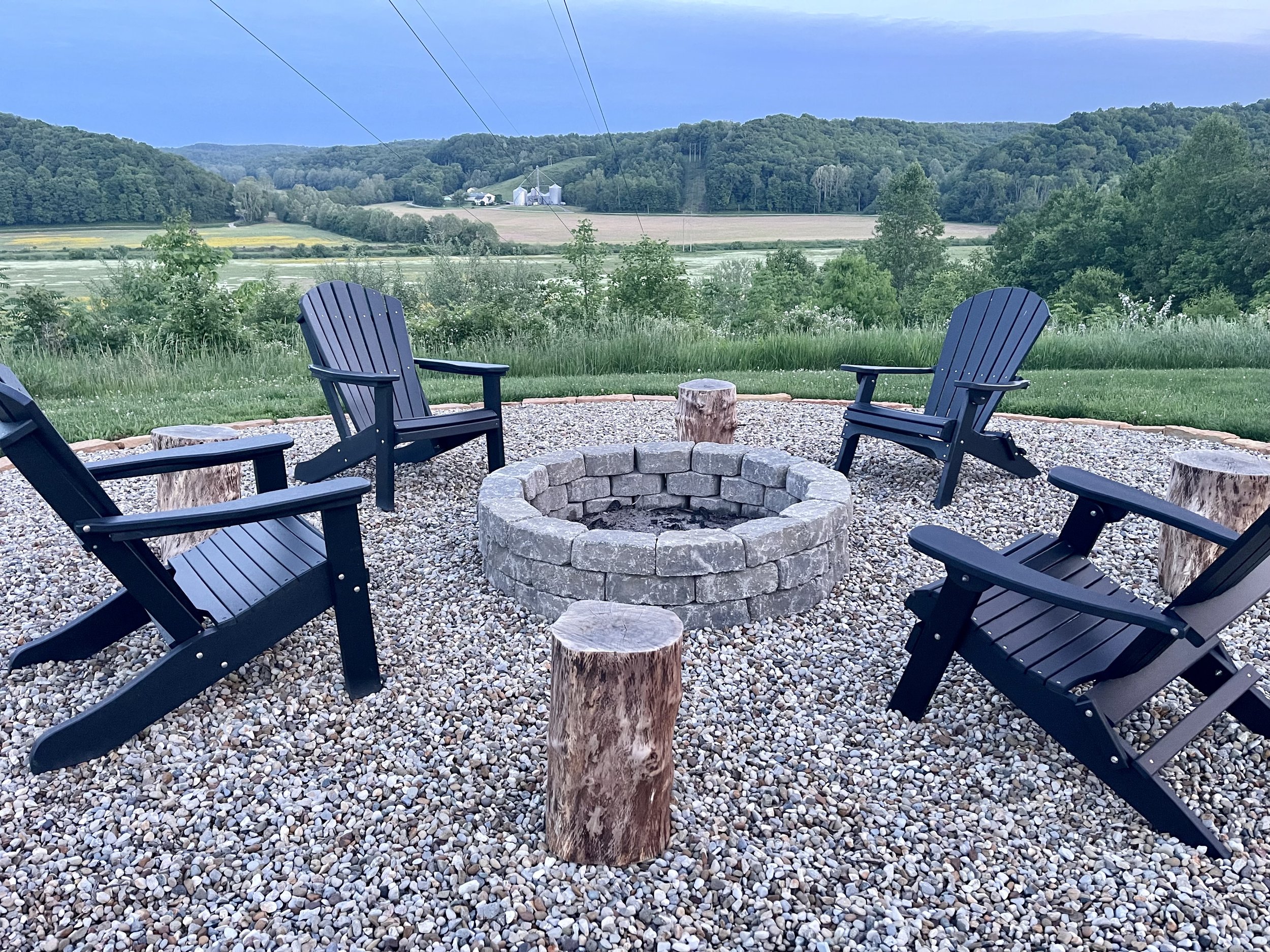 Outdoor fire pit area with four black Adirondack chairs and wooden stools on gravel, overlooking green hills and farms under a blue sky.