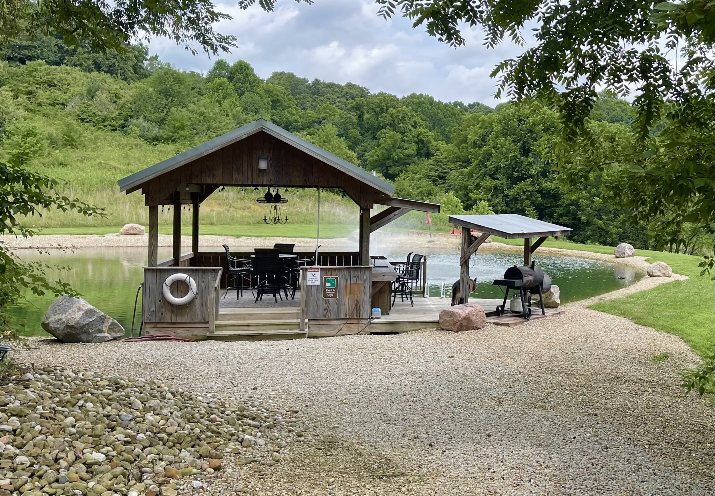 A lakeside wooden pavilion with a roof, surrounded by a gravel pathway, rocks, and lush green trees in the background. There are tables, chairs, a grill, and a life preserver hanging on the pavilion.