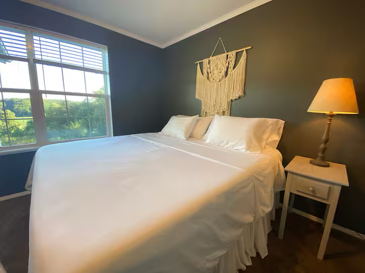 A bedroom with a large bed covered in white linens, a small white nightstand with a lamp, and a window showing a green outdoor view. There is a decorative wall hanging above the bed.