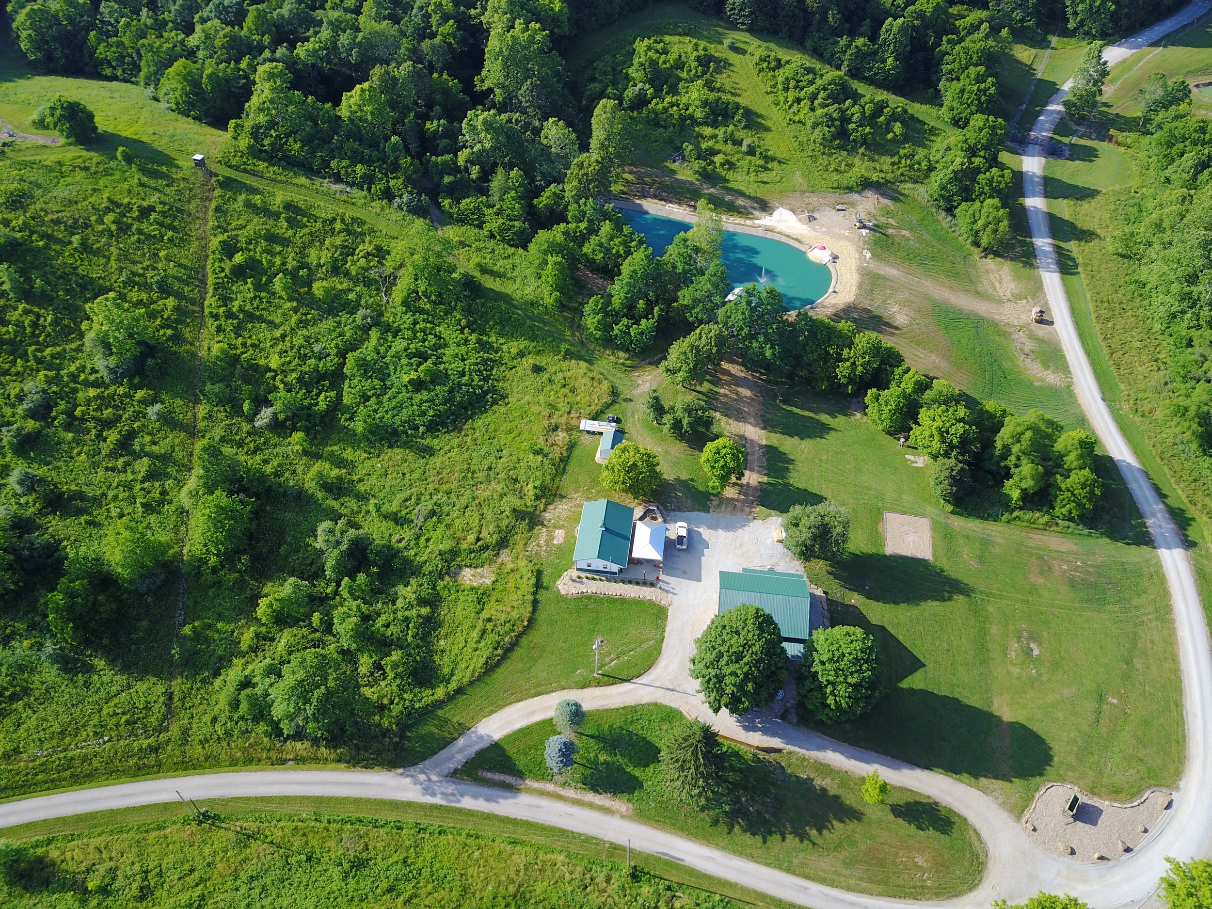 Aerial view of a rural landscape with a pond, trees, and several buildings, including a driveway and parked vehicles.