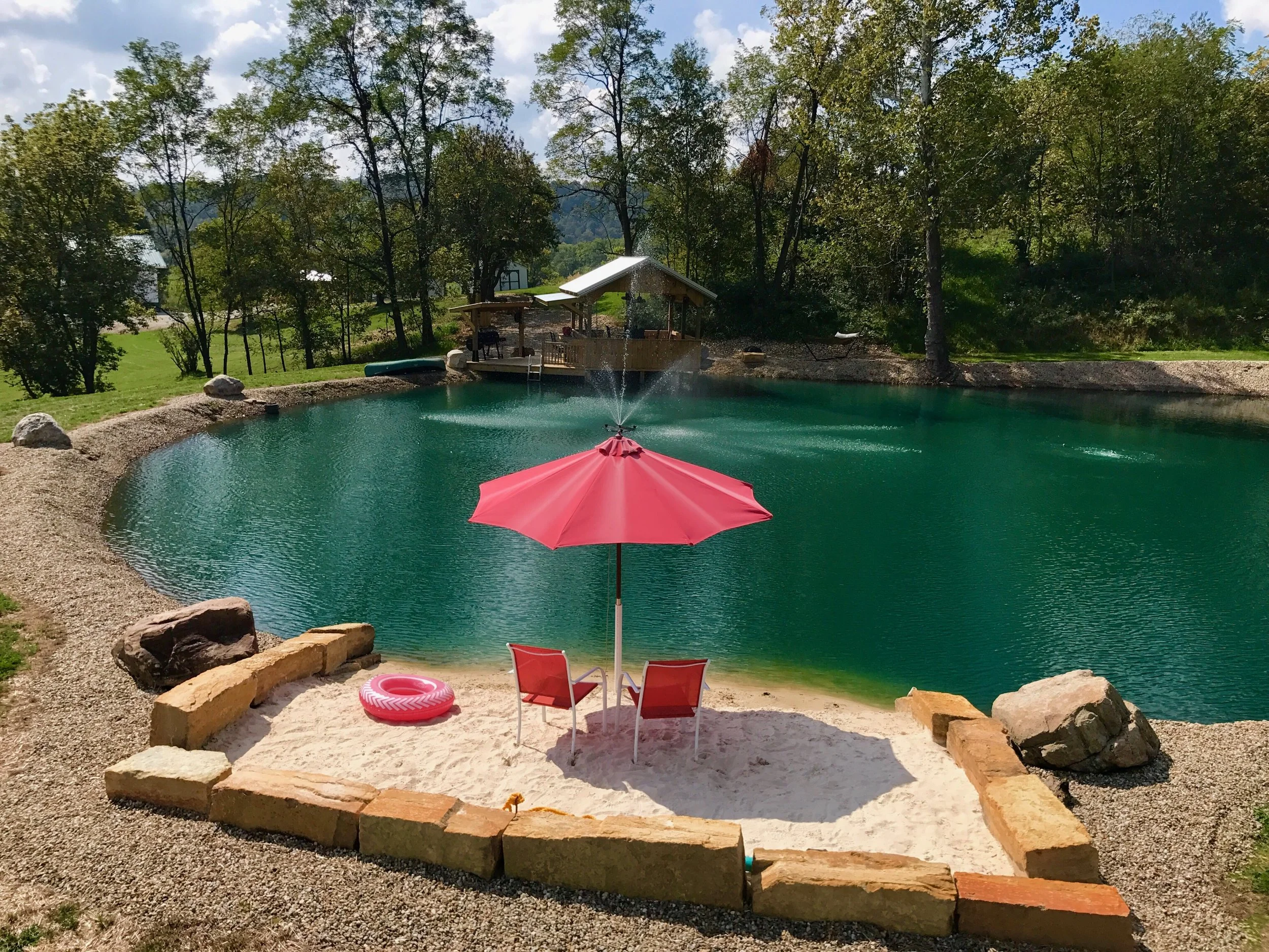 A backyard with a small pond that has a fountain in the middle, surrounded by trees. In the foreground, there's a sandy area with two red chairs, a pink umbrella, a pink inflatable ring, and a stone border. Behind the pond, there is a wooden structur