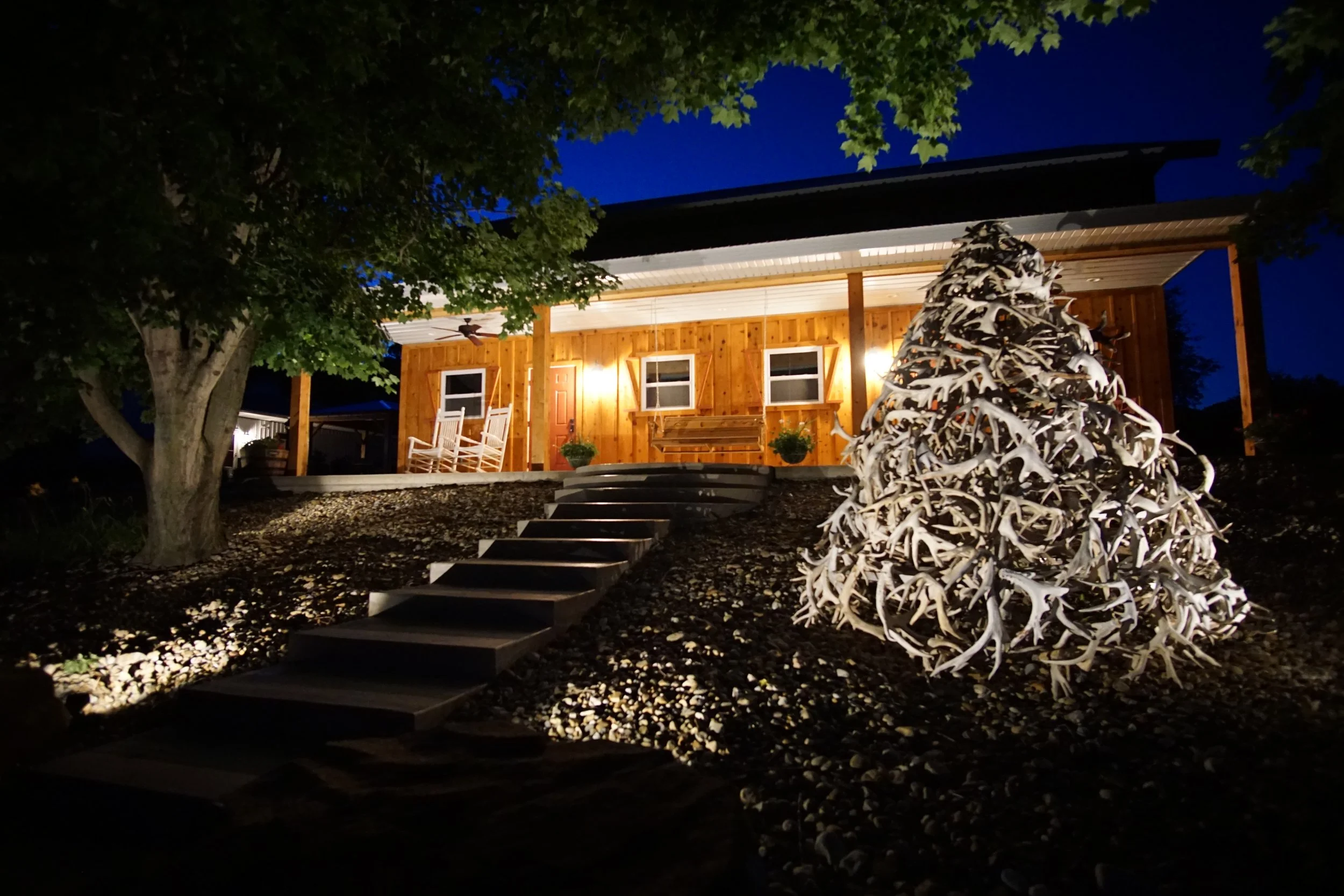 A wooden house with a lit porch, surrounded by trees and landscaping, including a whitetail deer antler-covered decorative tree sculpture in the yard at night.