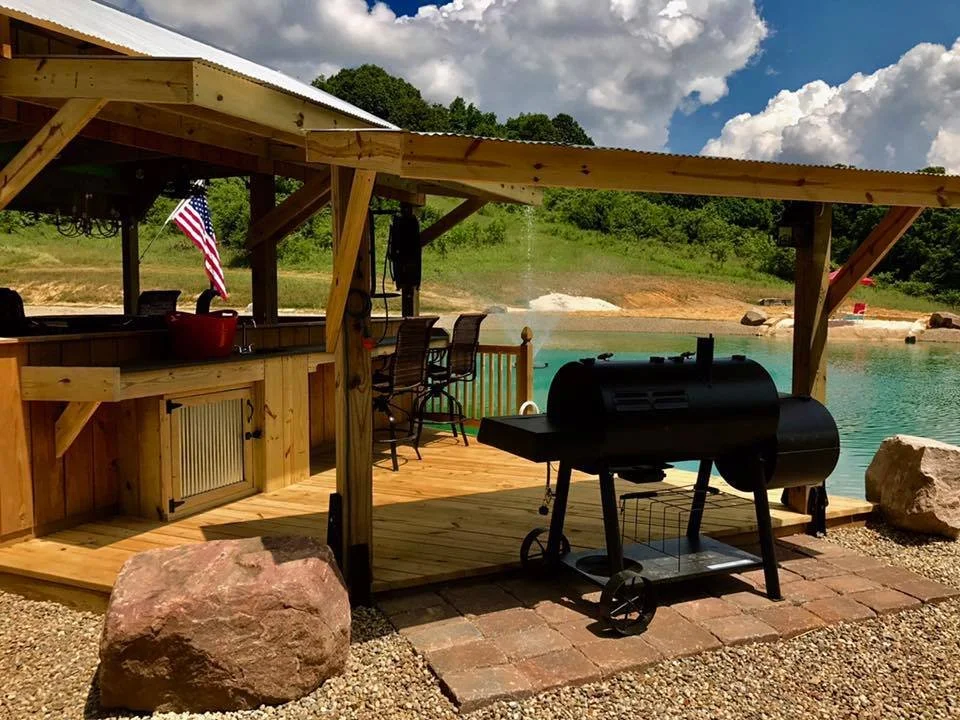 A wooden outdoor bar with a barstool, a portable grill, and an American flag, overlooking a lake with a sandy shoreline and green trees in the background.