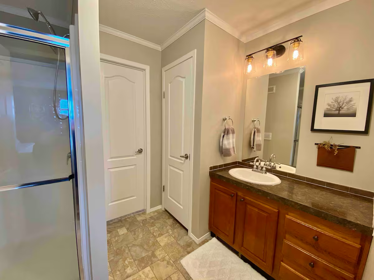 Bathroom with walk-in shower, wooden vanity with dark countertop, large mirror, framed black-and-white tree photo, gray walls, tiled floor, and lighting fixture above the mirror.