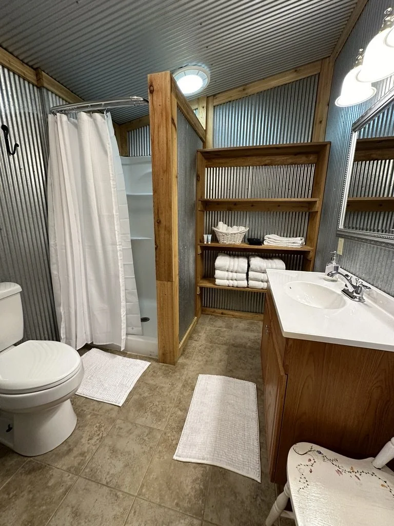 A rustic bathroom with corrugated metal walls, a wooden ceiling, and a tiled floor. It features a white toilet, a shower with a white curtain, a wooden shelving unit with folded towels, a white countertop with a sink, a mirror, and a small chair.