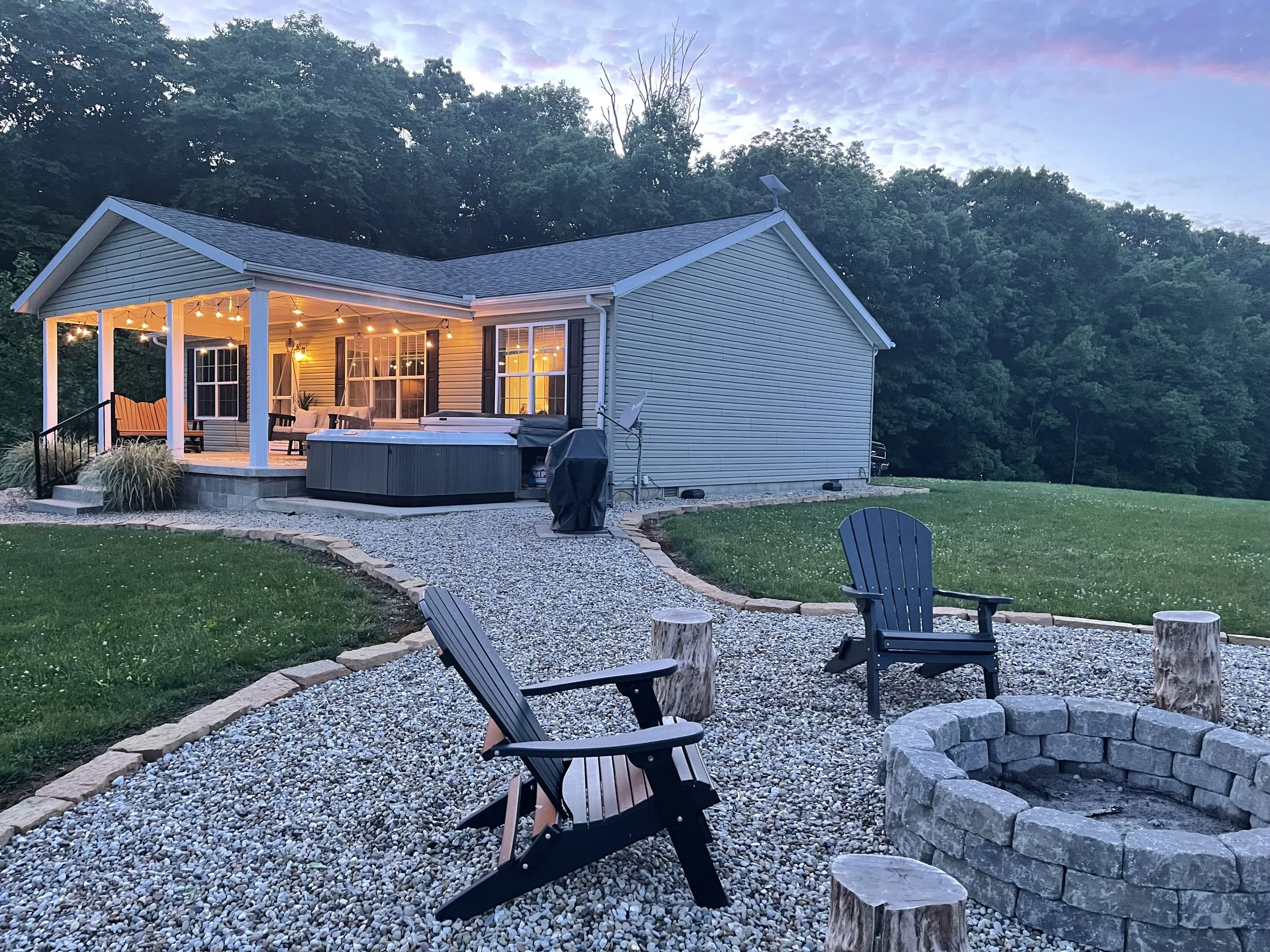 A backyard scene with a house featuring a lit porch, a hot tub, two Adirondack chairs, and a fire pit with tree stumps, surrounded by green grass and trees in the background at dusk.