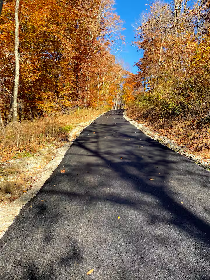 A freshly paved asphalt road winds through a wooded area with colorful autumn foliage, under a bright blue sky with a few clouds.