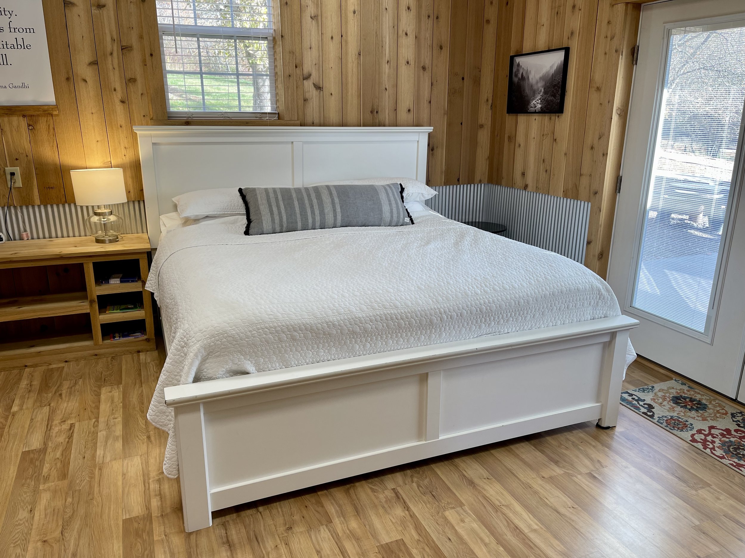 A cozy bedroom with wooden walls, featuring a white bed with a white quilt and a gray bolster pillow. Next to the bed, there's a wooden nightstand with a lamp, books, and a phone. A window and a glass door with blinds let in natural light, and a smal