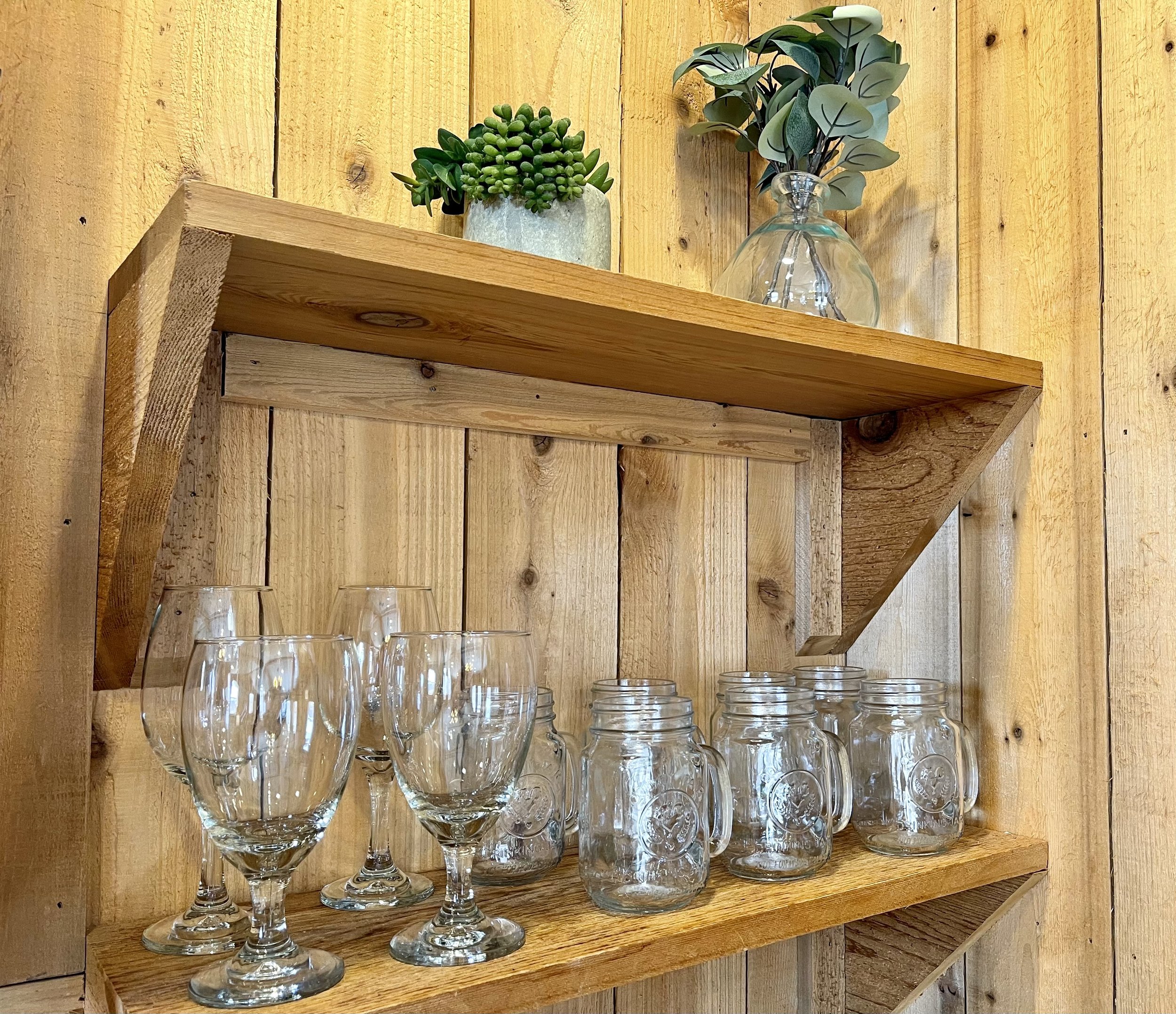 Wooden wall shelf holding potted plants and glassware, with a background of vertically arranged wooden planks.