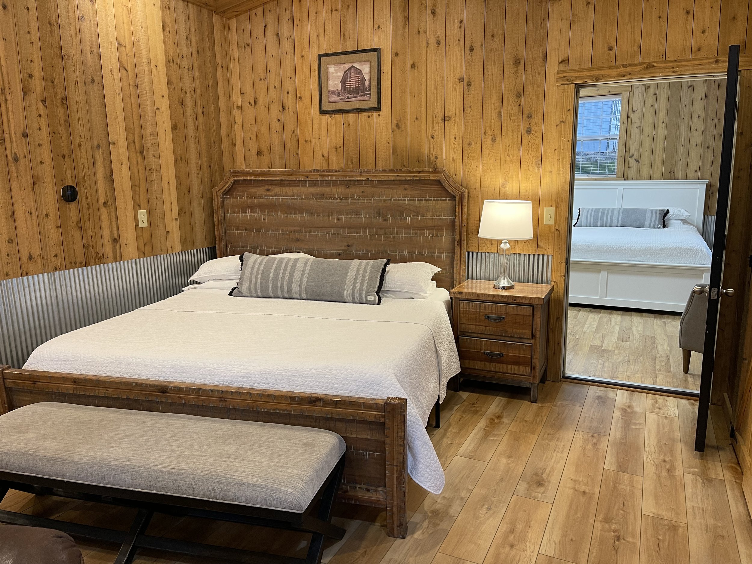 A cozy bedroom with wooden walls, a large bed with white bedding and a striped bolster pillow, a wooden nightstand with a lamp, and a mirror reflecting an adjacent room with another bed.