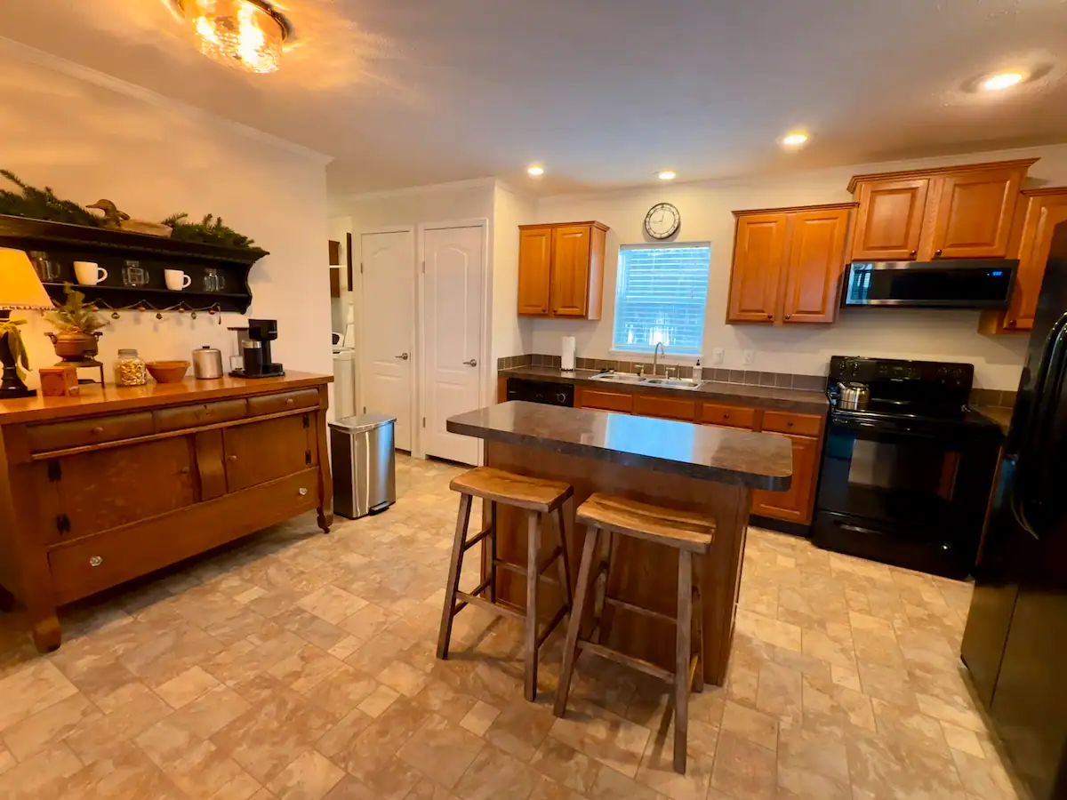 Kitchen with wooden cabinets, a small island with two wooden stools, black appliances, and a decorated shelf on the wall.