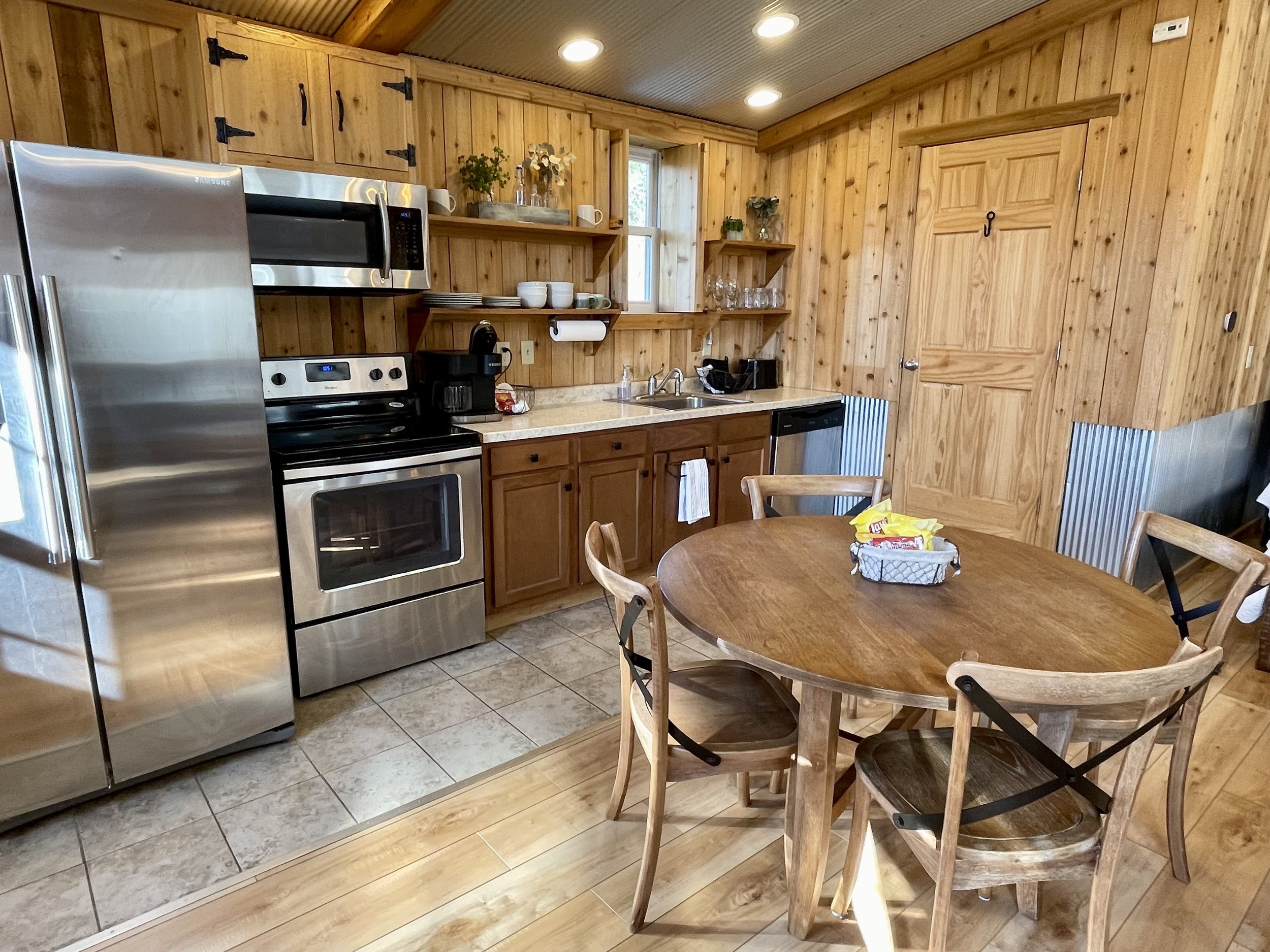 Kitchen with wood-paneled walls, stainless steel refrigerator, microwave, stove, open wooden shelves with dishes and glassware, round wooden dining table with four chairs, and kitchen essentials like coffee maker, sink, and paper towel holder.