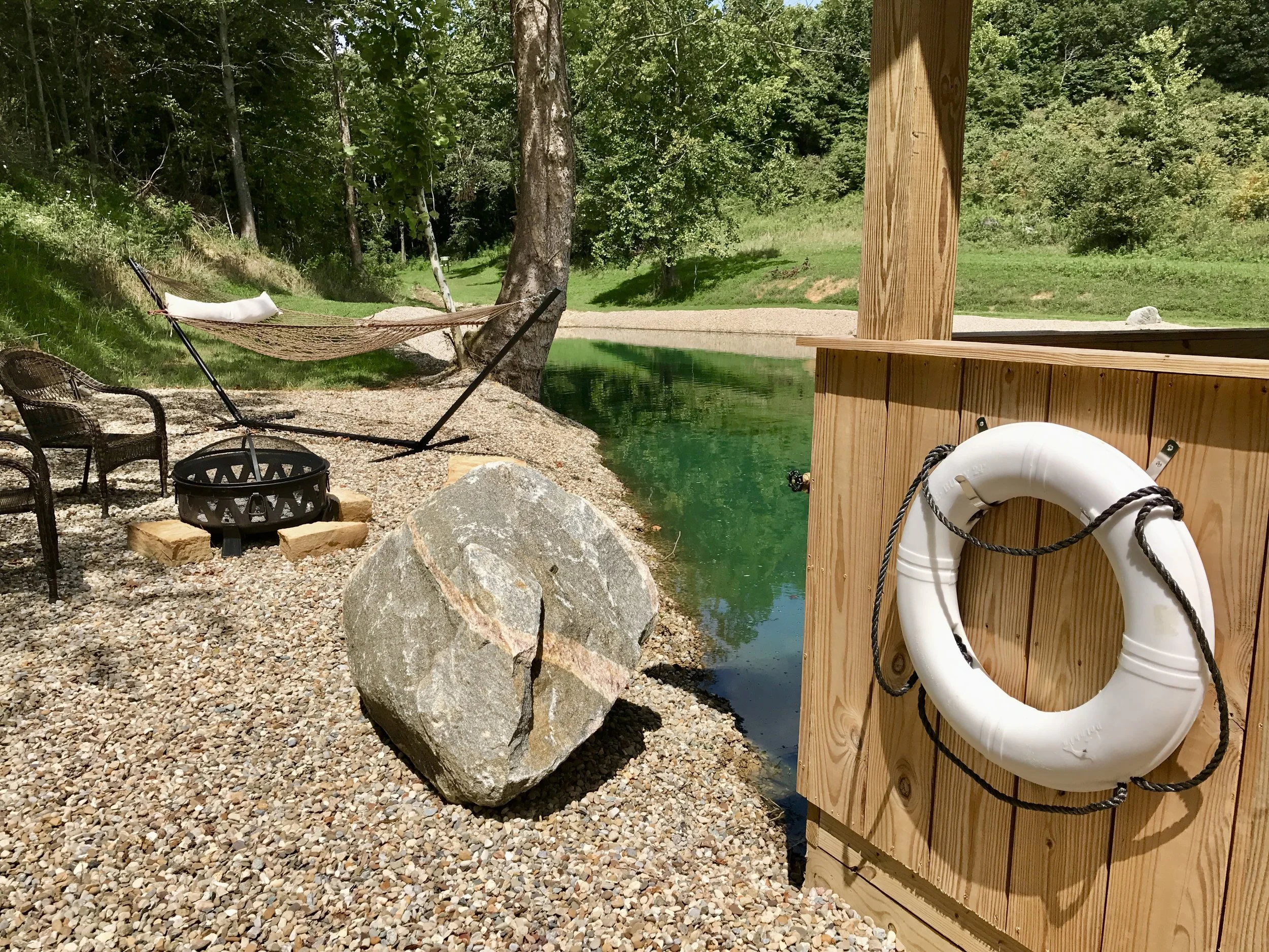 Outdoor lakeside scene with a picnic table, a white life ring mounted on a wooden structure, a black fire pit, rocks, gravel ground, a hammock between trees, and a view of green trees and water.
