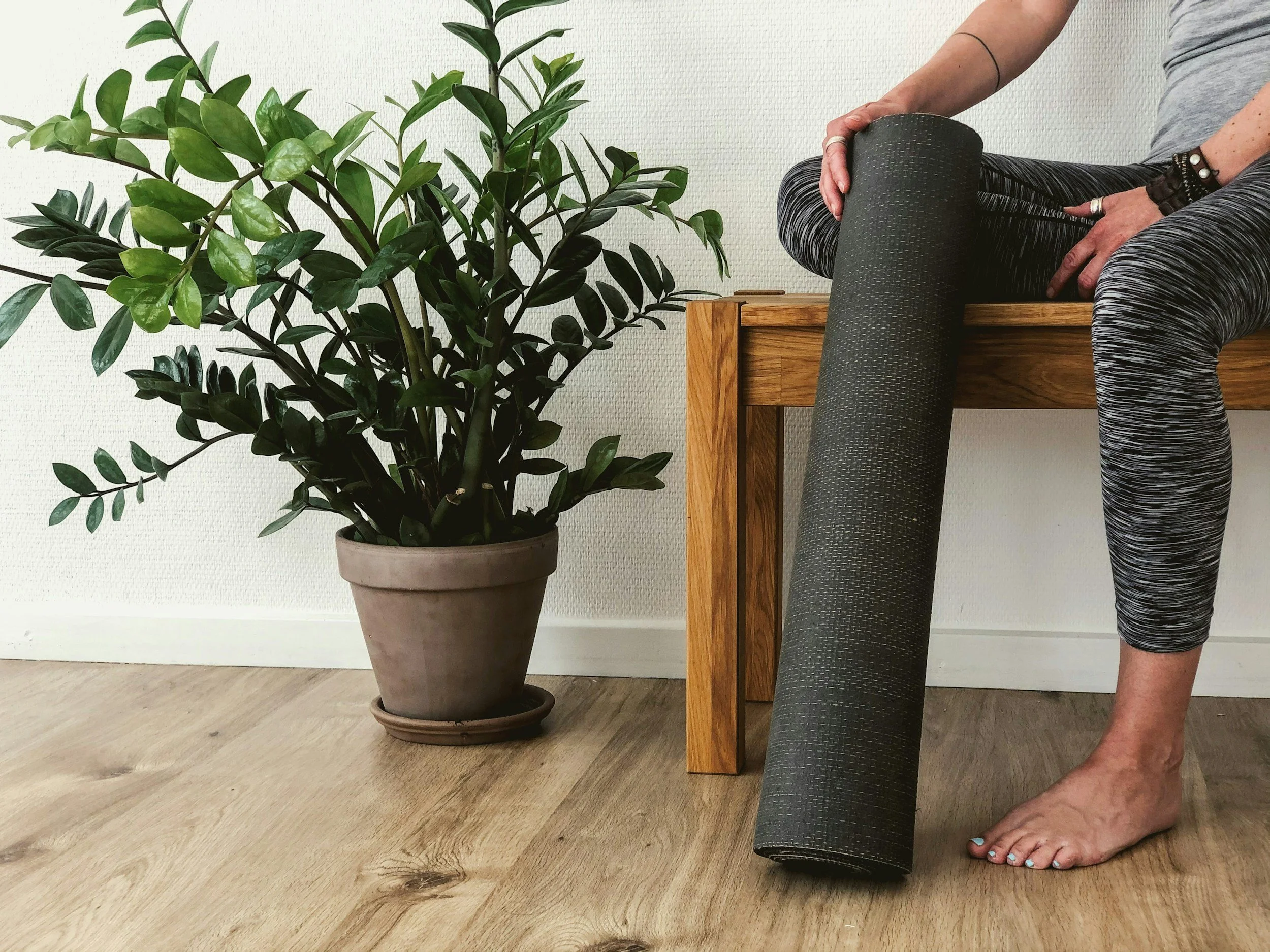 Person sitting on a wooden bench holding a rolled-up yoga mat, with a large potted plant nearby in a room with wooden floor and white wall.