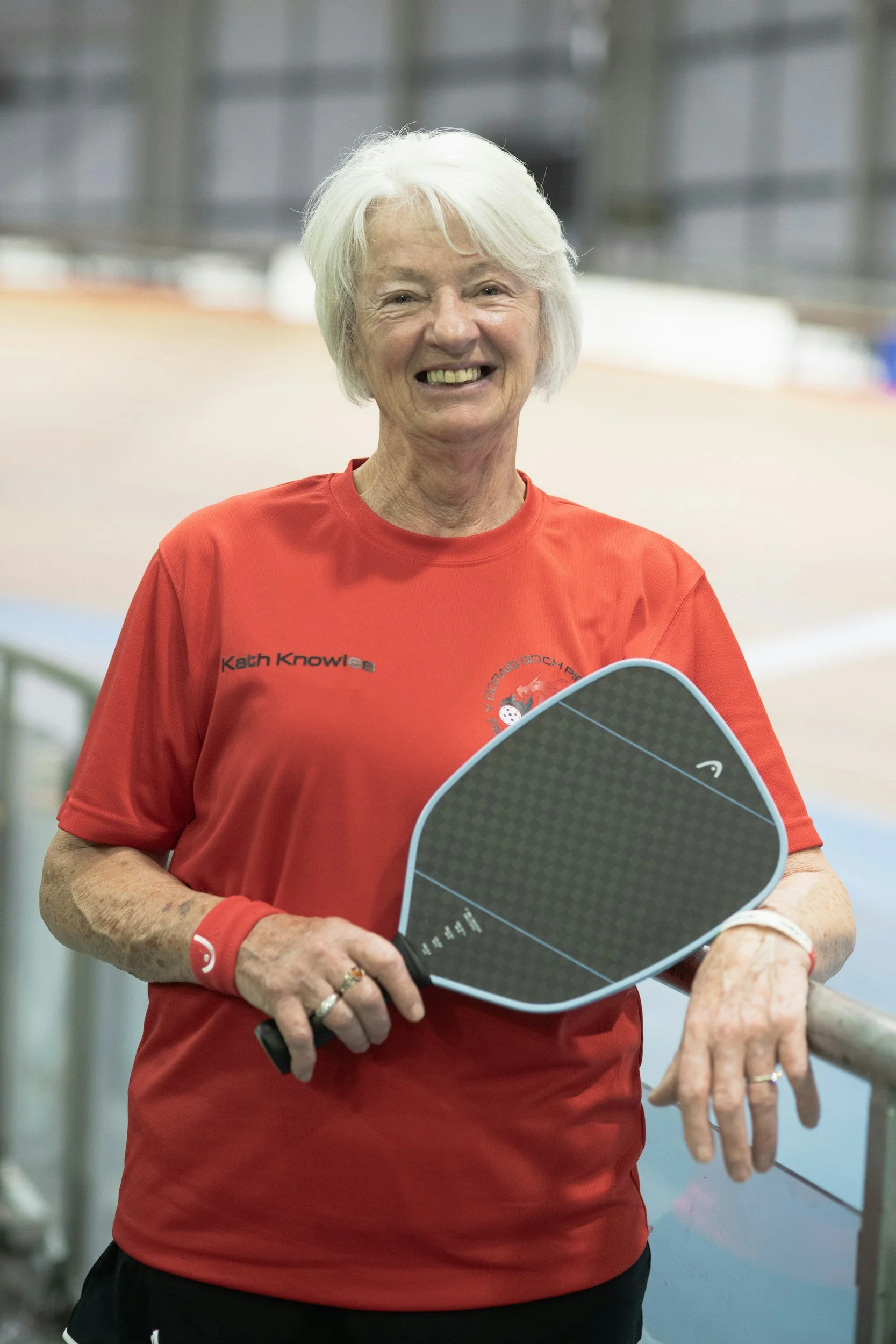 An elderly woman smiling and holding a paddle, wearing a red sports shirt, standing indoors at a sports facility.
