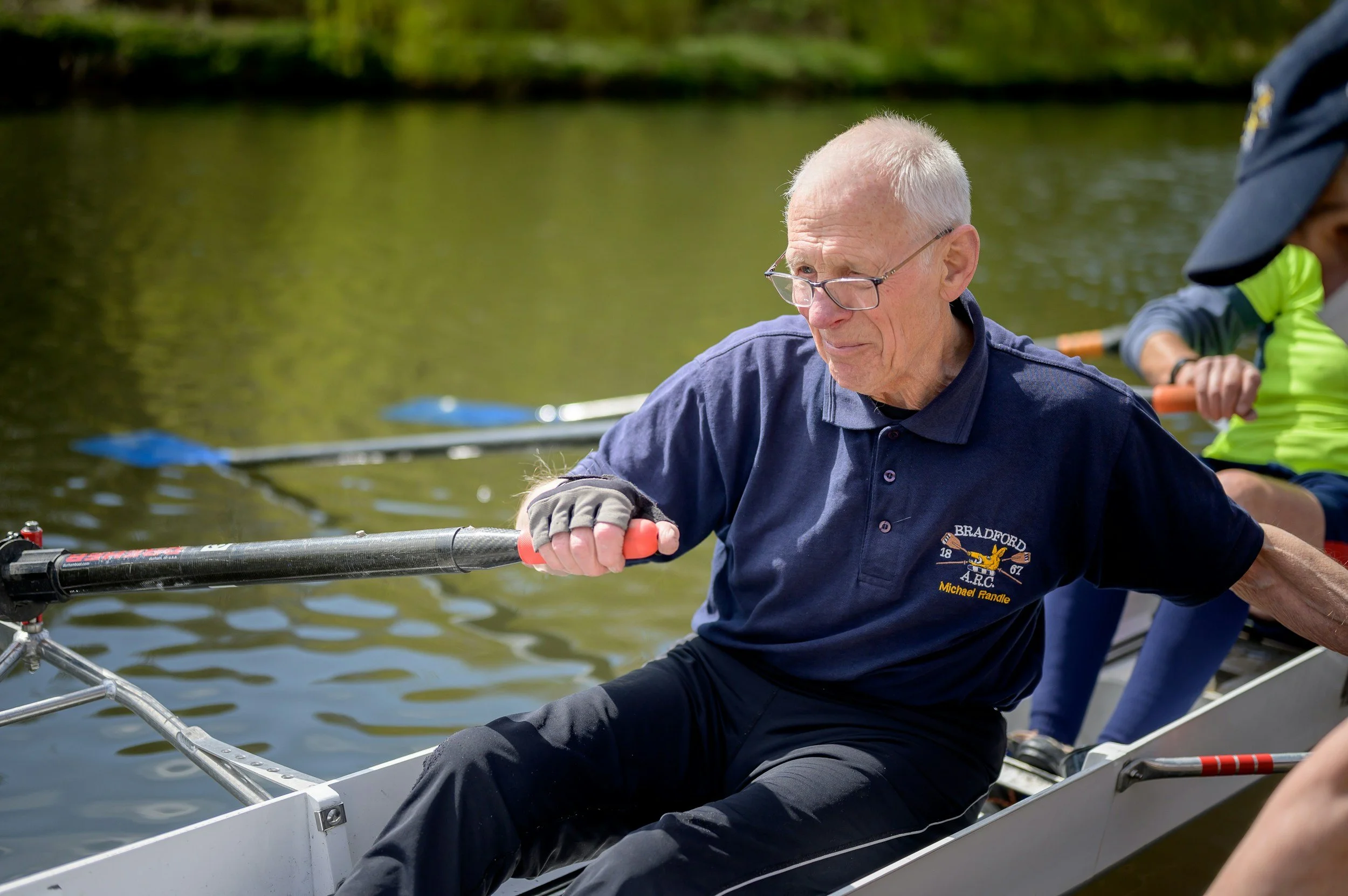 Elderly man rowing a boat on a calm body of water, wearing glasses and a navy blue polo shirt with a logo.
