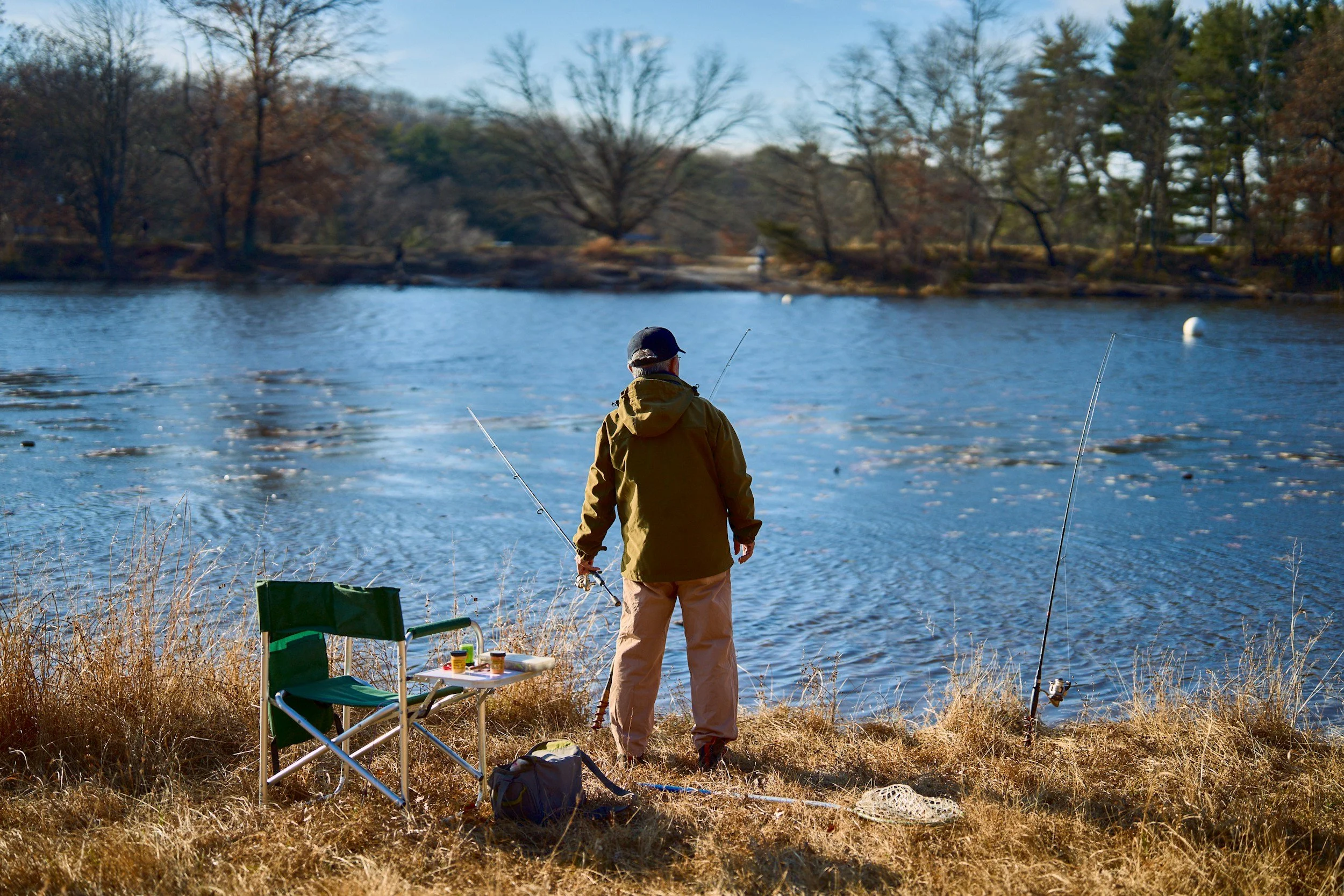 Person fishing by a lake with fishing rods, a green camping chair, and a small table with supplies, during daytime in a natural outdoor setting.