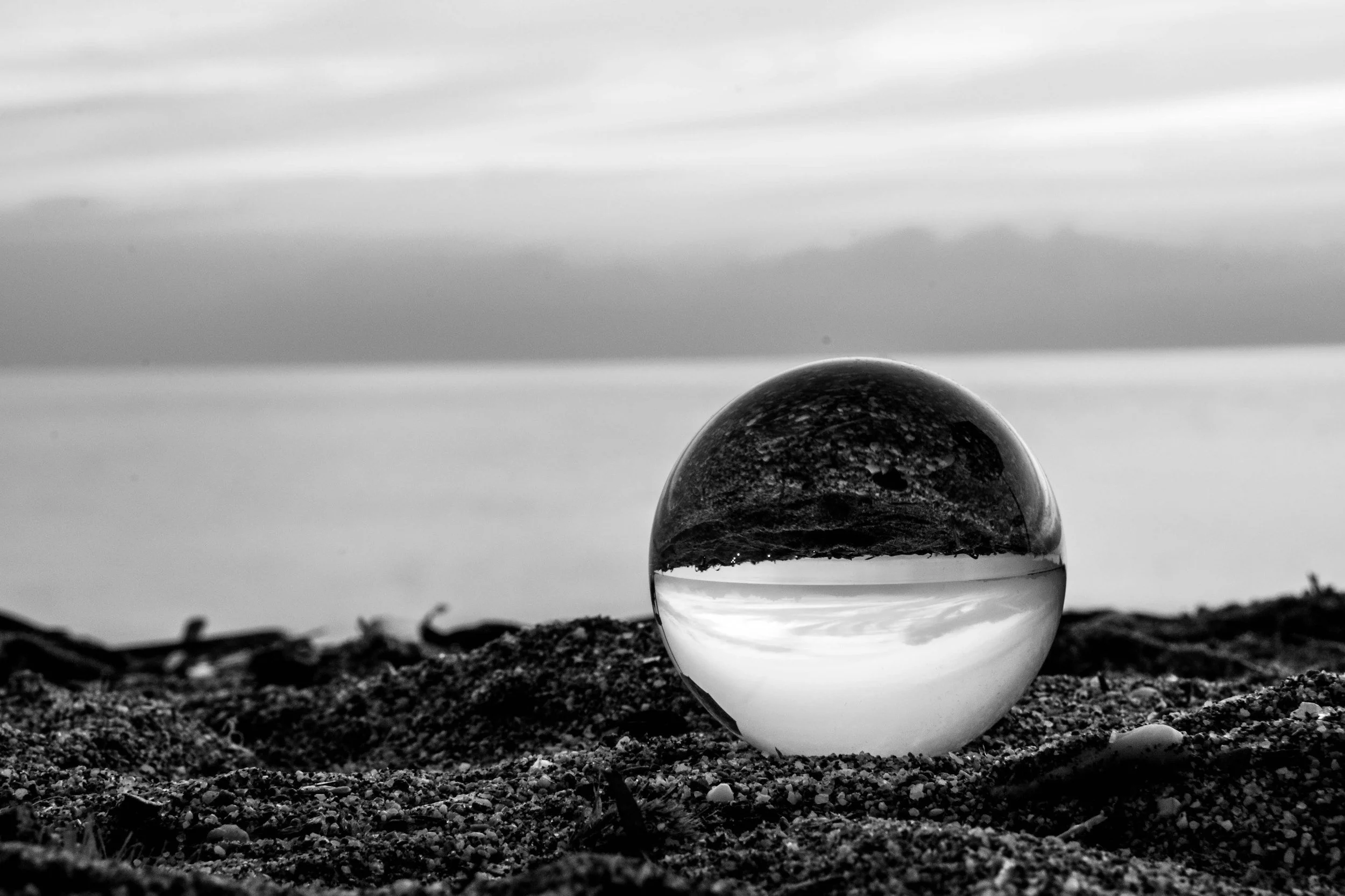 A clear glass sphere placed on the ground, reflecting the cloudy sky and surrounding landscape in black and white.