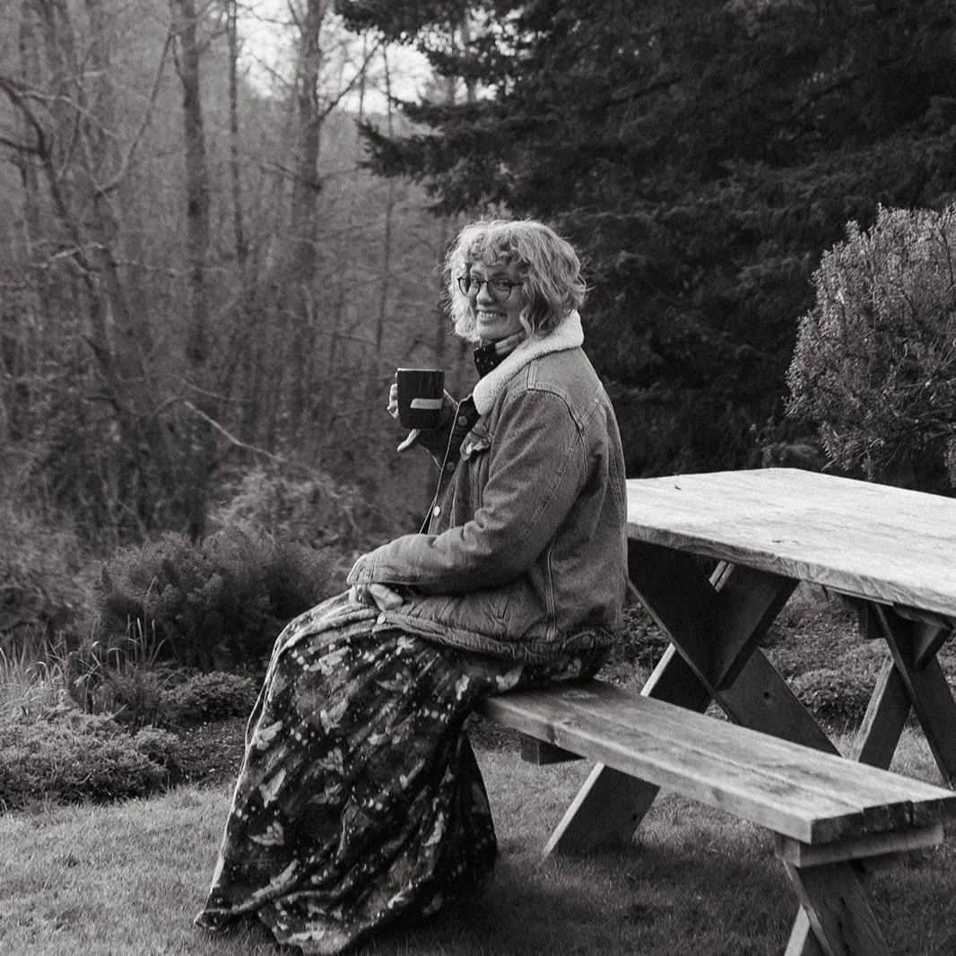 A woman with curly hair and glasses sitting on a wooden bench outdoors, holding a mug, in a natural setting with trees and bushes, captured in black and white.