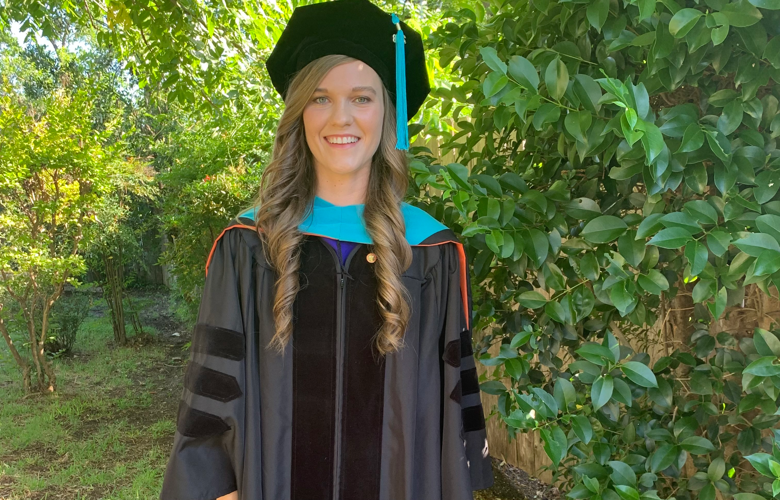 Young woman in graduation cap and gown standing outdoors among green foliage, smiling, during daytime.