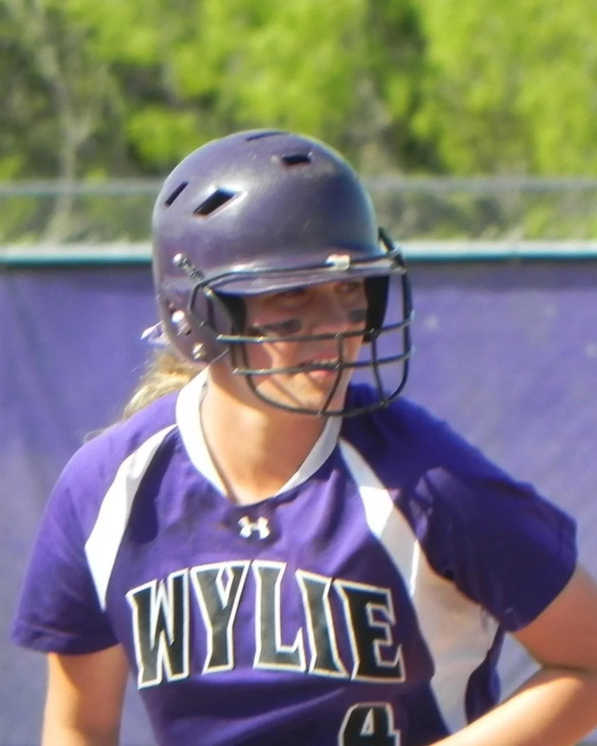 Female softball player wearing a purple jersey with white accents and a black helmet, standing outdoors on a field.
