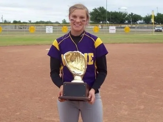 A young female softball player in purple and yellow uniform holding a trophy on a softball field.